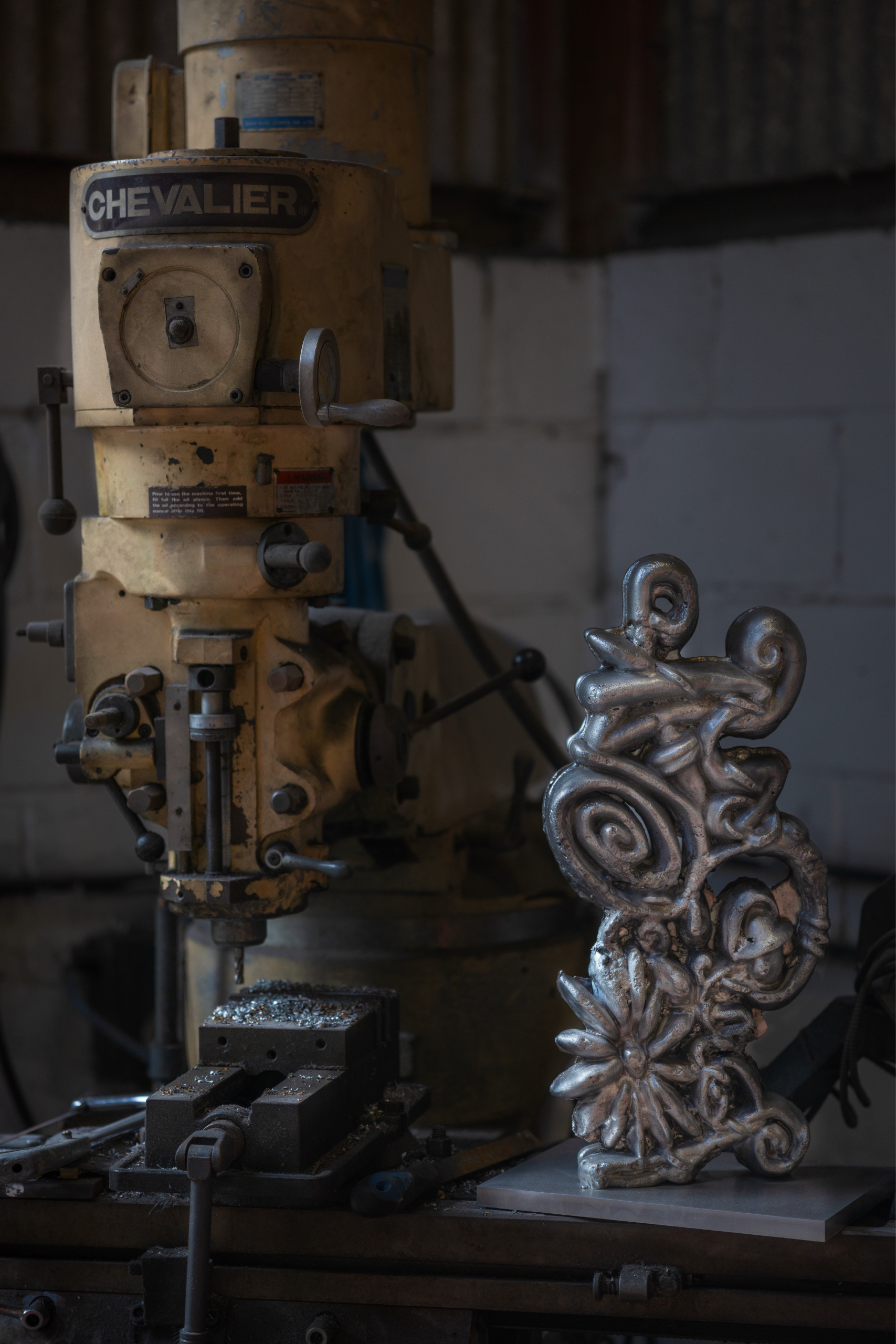 A metal sculpture with intricate swirling designs and floral elements, standing on a base next to a yellow industrial machine in a workshop.