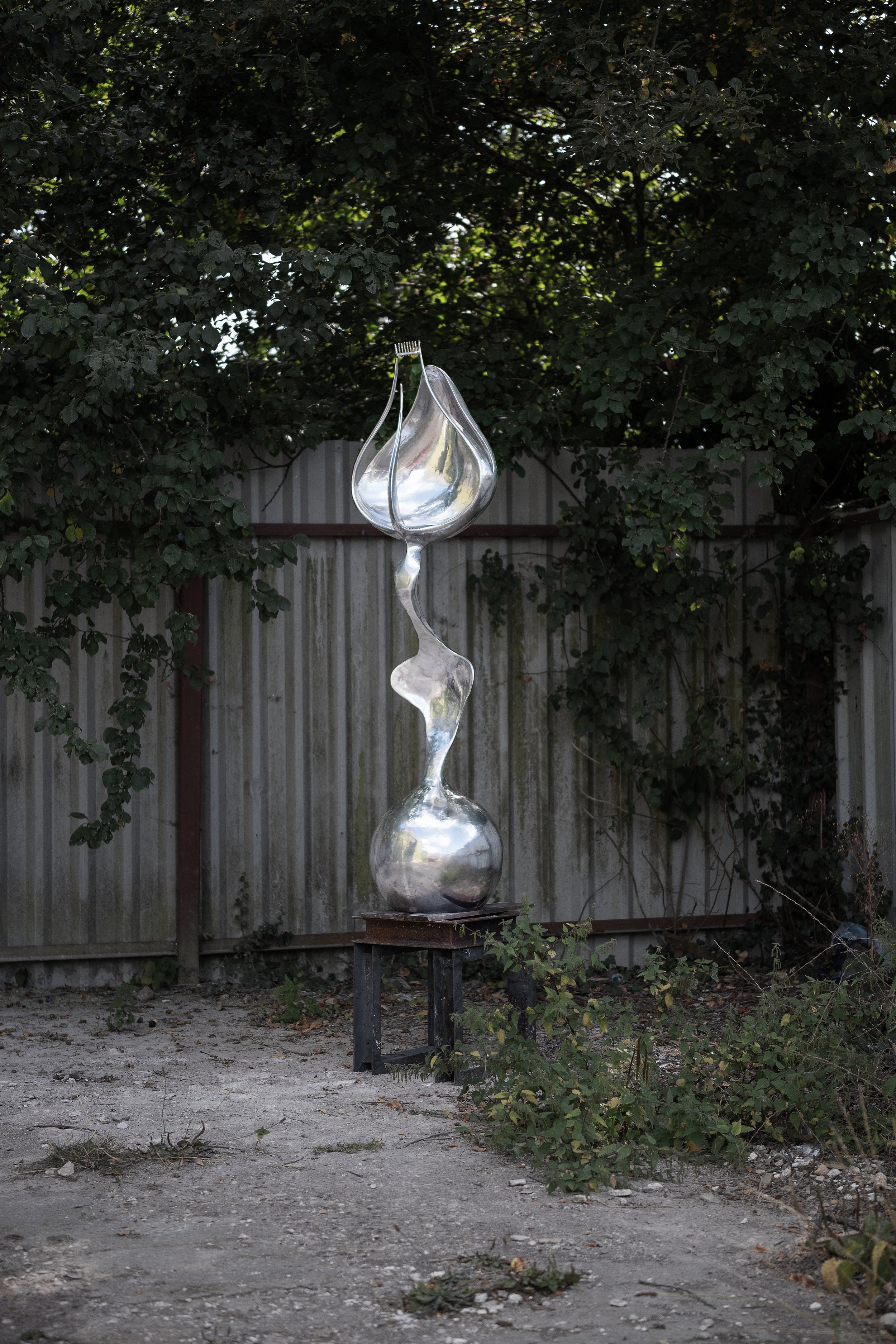 Sculpture made of polished stainless steel, shaped like interconnected teardrops or abstract forms, outdoors against a background of trees and a fence.