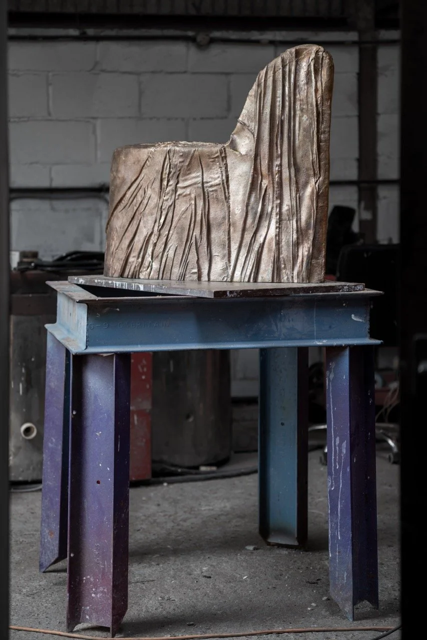 A metal sculpture of a cliff or mountain made of bronze or similar material, placed on a metal stand in a workshop.