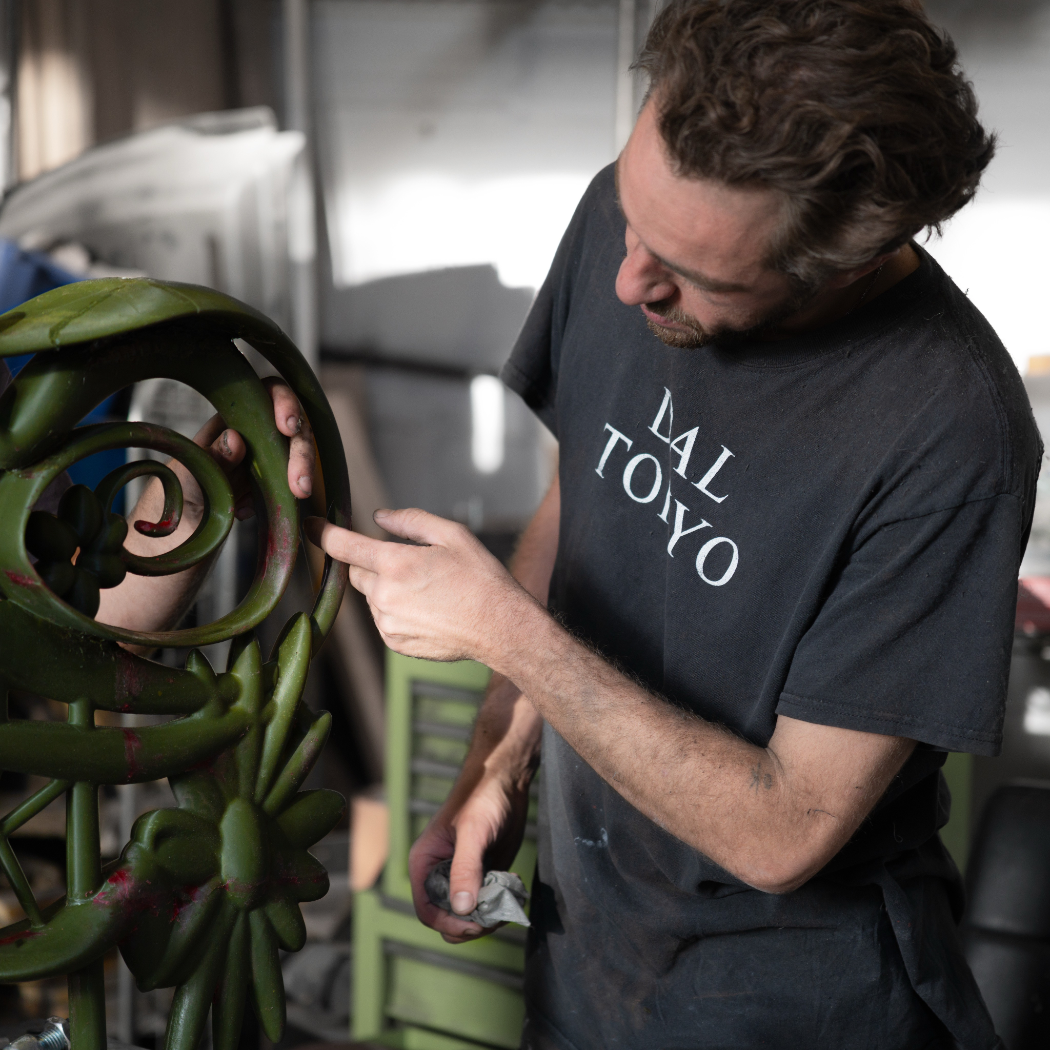 A man working on a green metal sculpture in a workshop, pointing at a specific part with his finger.