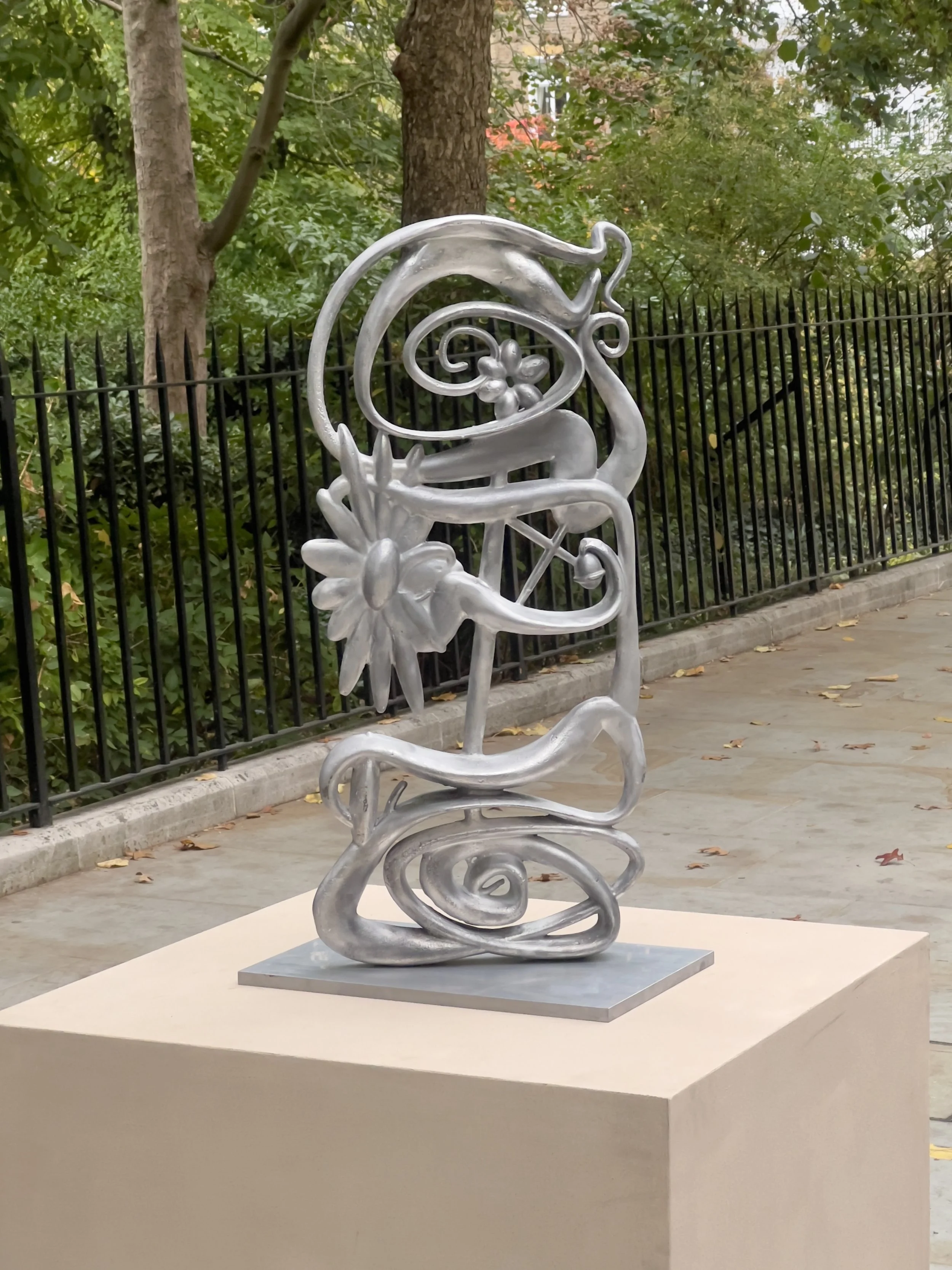 Silver abstract sculpture with flowing curves, floral motifs, and a snail, displayed on a pedestal outdoors.