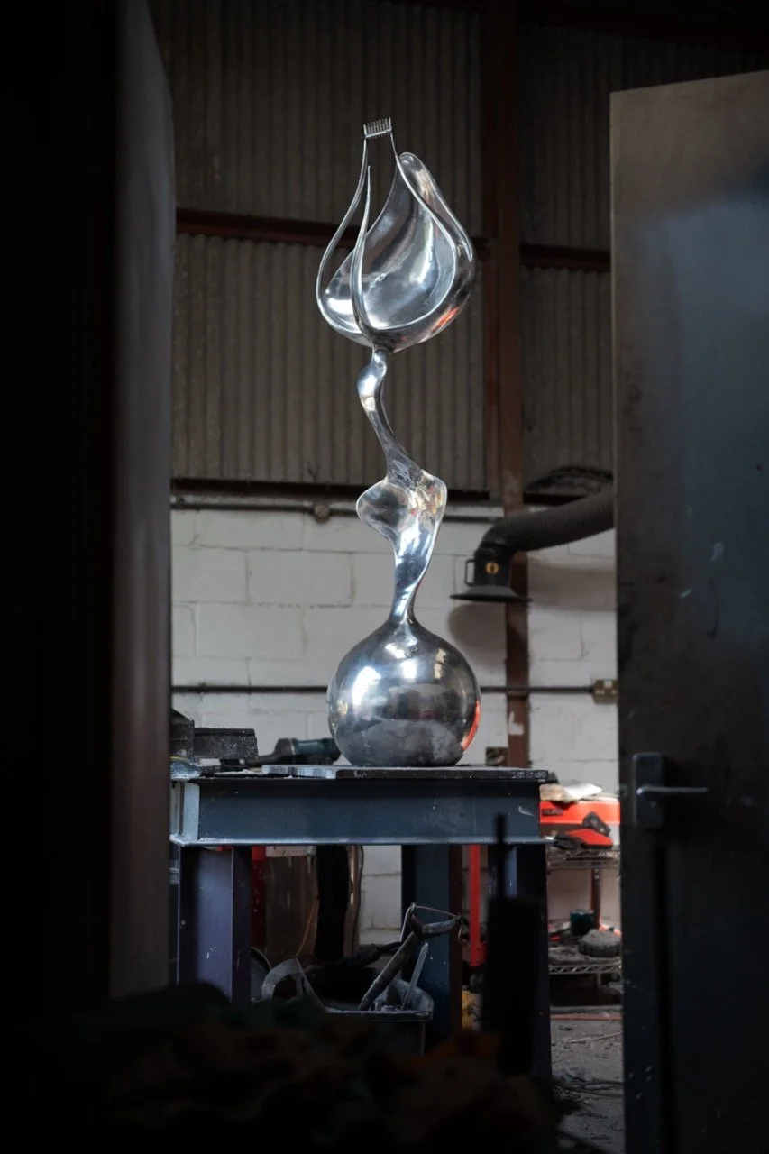 A metallic abstract sculpture on a black table in an industrial workshop with a corrugated metal ceiling and a white brick wall in the background.