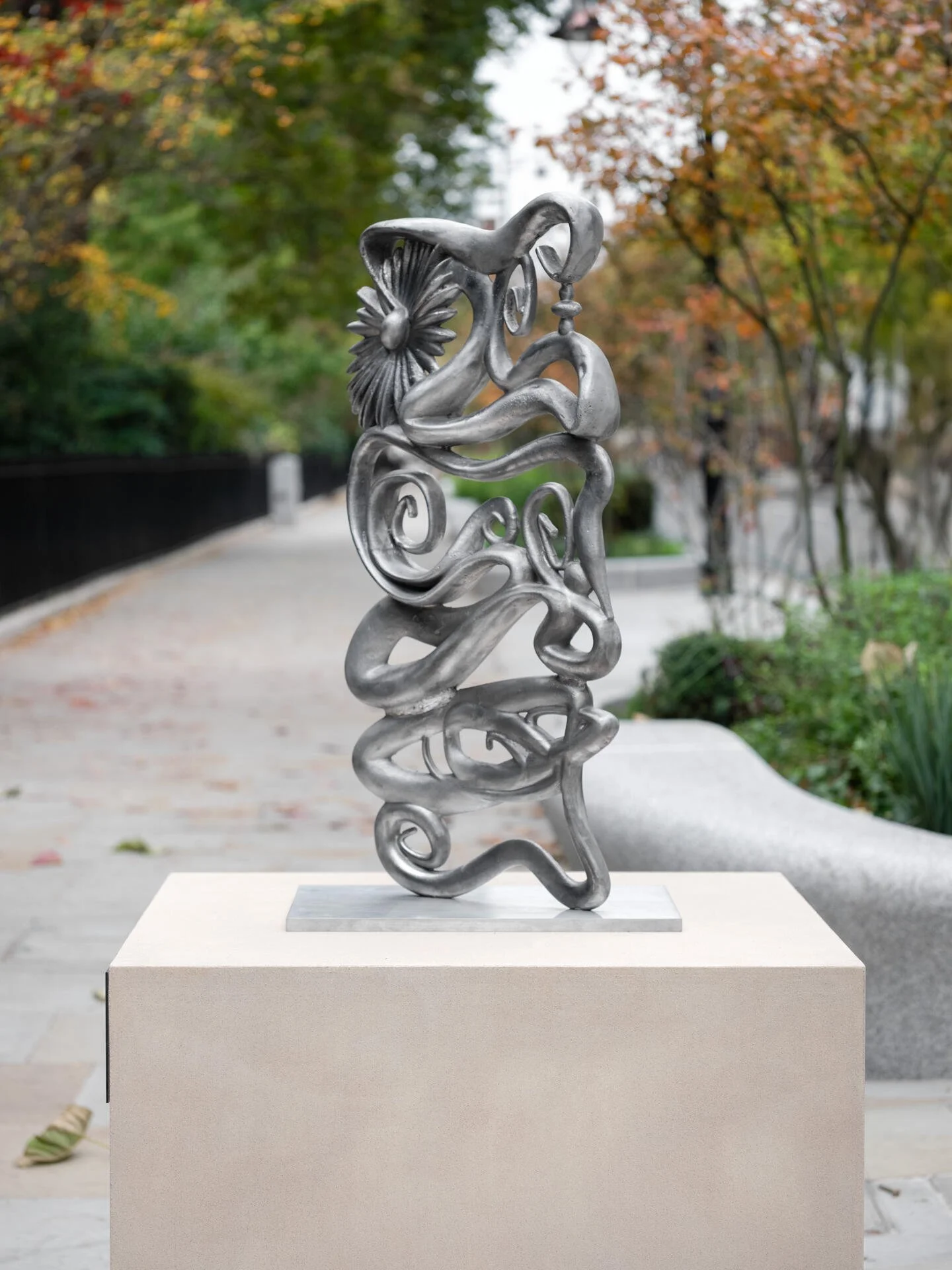 Abstract metal sculpture with swirling shapes and a flower, displayed outdoors on a pedestal amid trees with autumn-colored leaves.