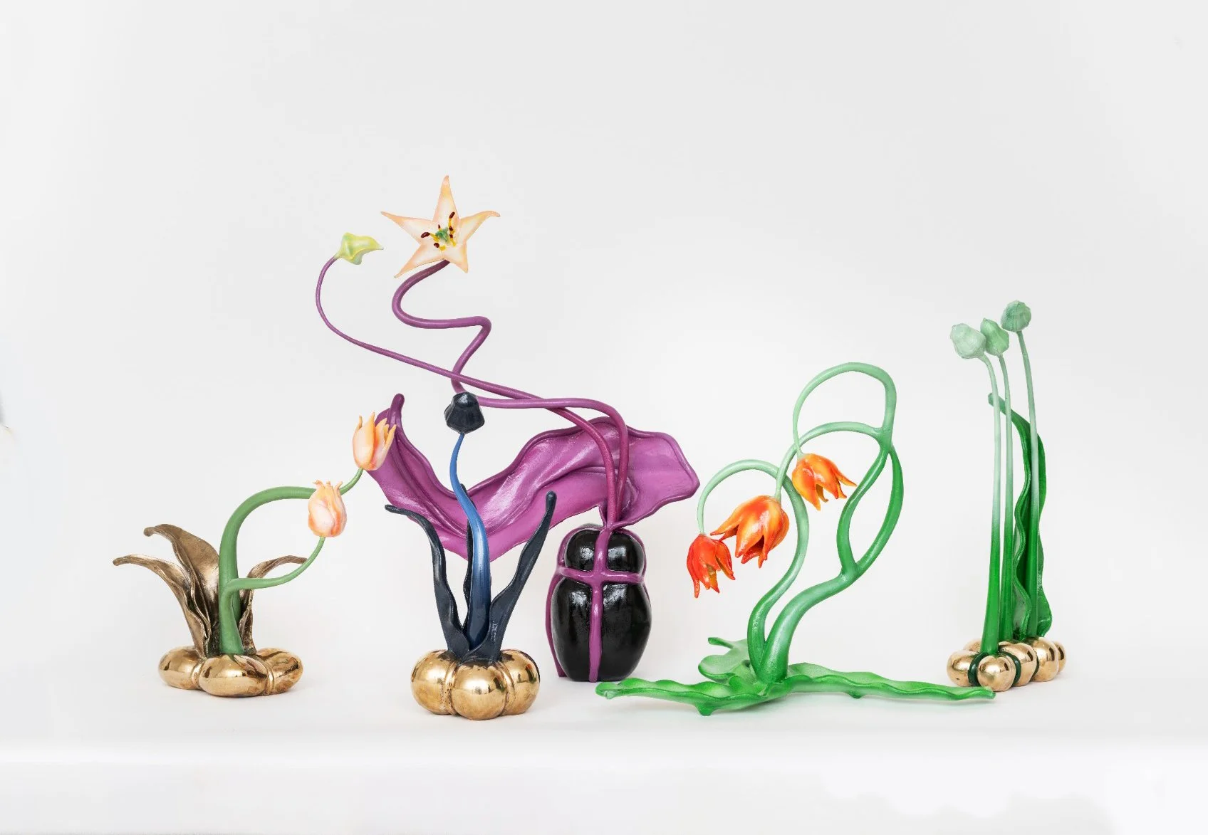 Colorful glass sculpture of various flowers, with a purple flower at the center, orange and pink flowers on either side, and a star-shaped flower at the top, all set against a plain white background.