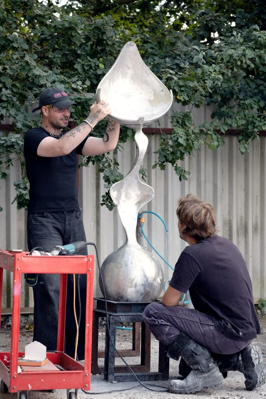 Two men work on a large, abstract metal sculpture outdoors. One man is holding the top of the sculpture, and the other is kneeling and observing. There are tools and equipment nearby.