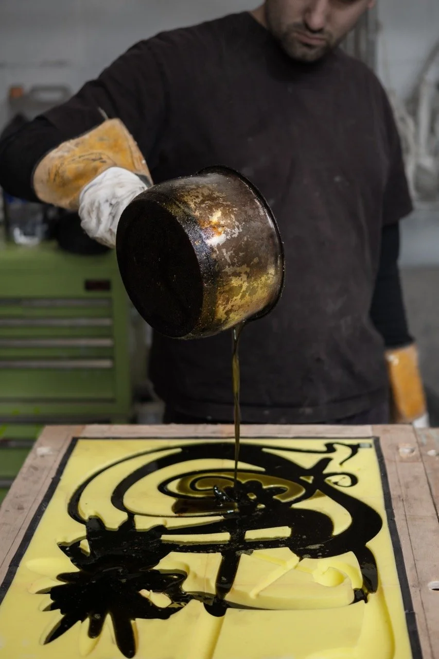 Person pouring black liquid paint onto a yellow canvas with abstract black design.