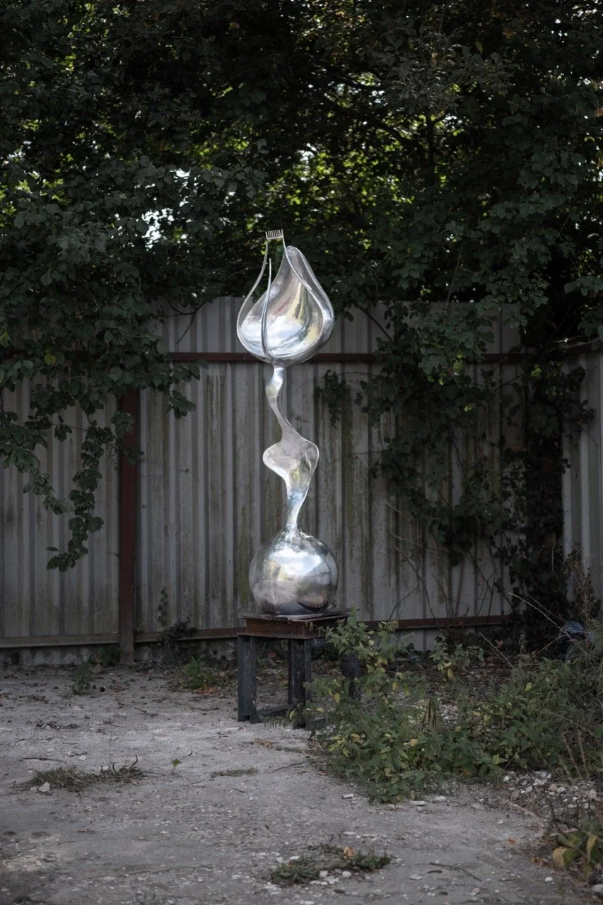 A tall abstract metal sculpture with a shiny, reflective surface is placed outdoors against a backdrop of trees and a corrugated metal fence. The sculpture has a curving, twisting form and rests on a small platform.