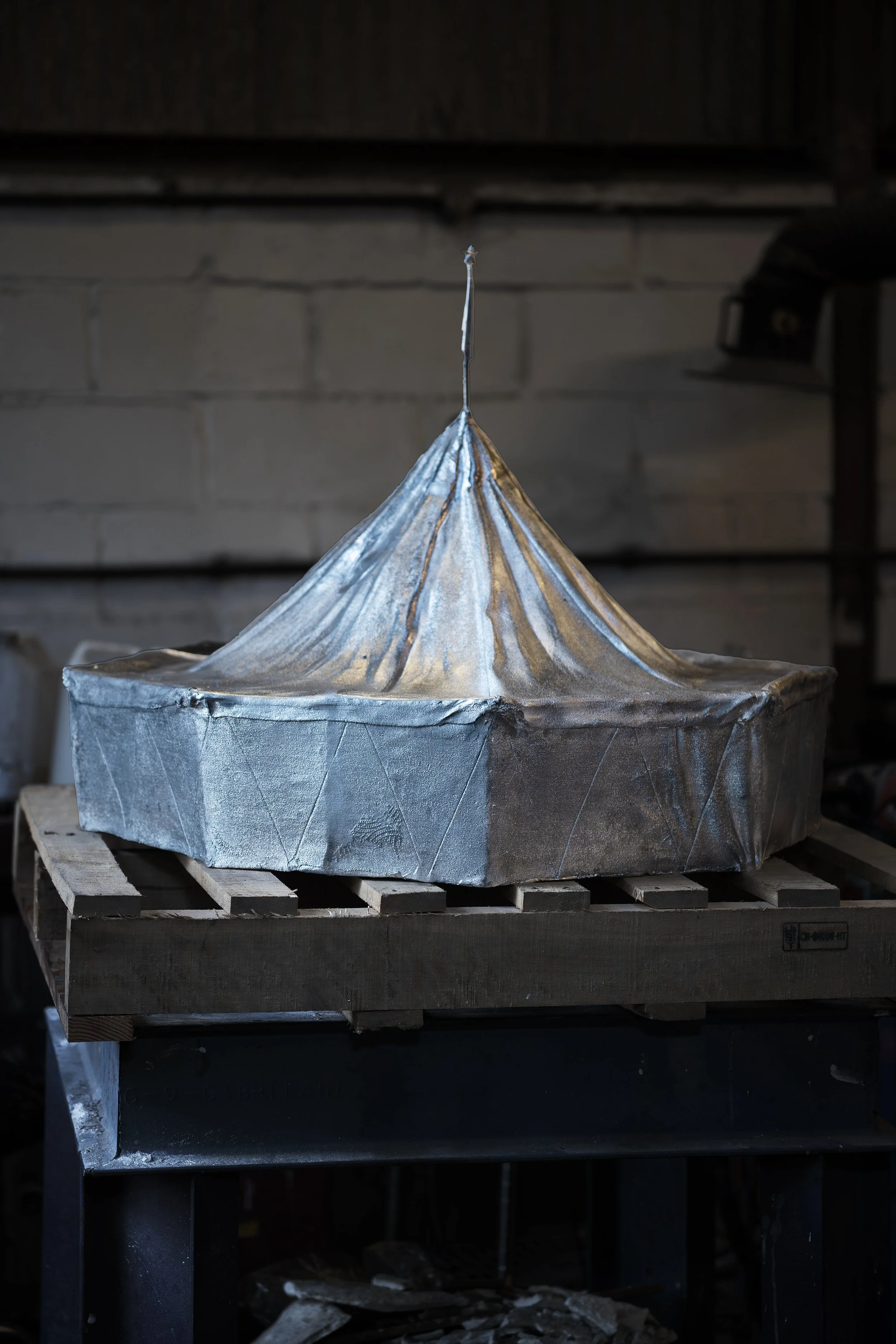 A metallic sculpture resembling a small house or castle with a conical roof, placed on a wooden pallet inside a workshop.