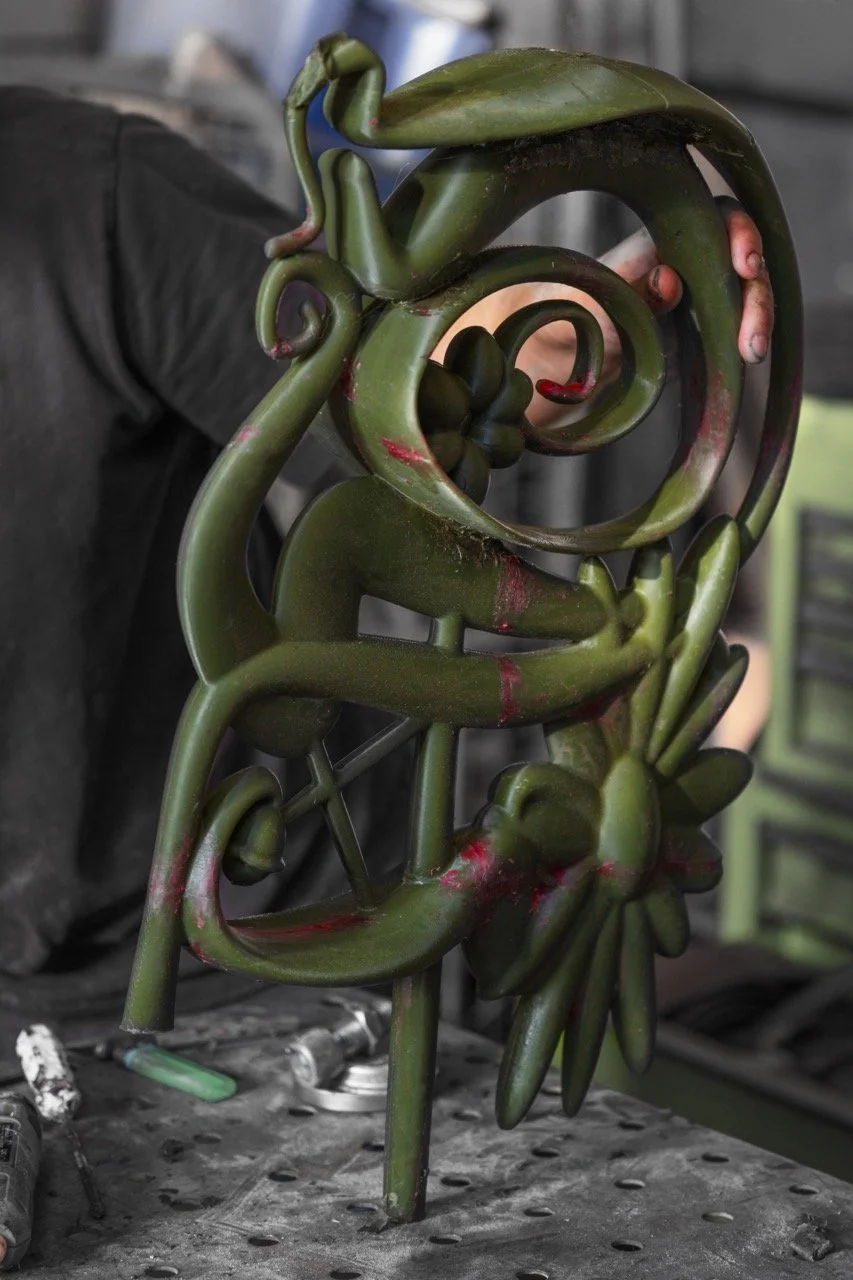 A person holding a green, twisted metal sculpture with spiral and floral designs. The sculpture has red paint streaks and is being held with one hand.