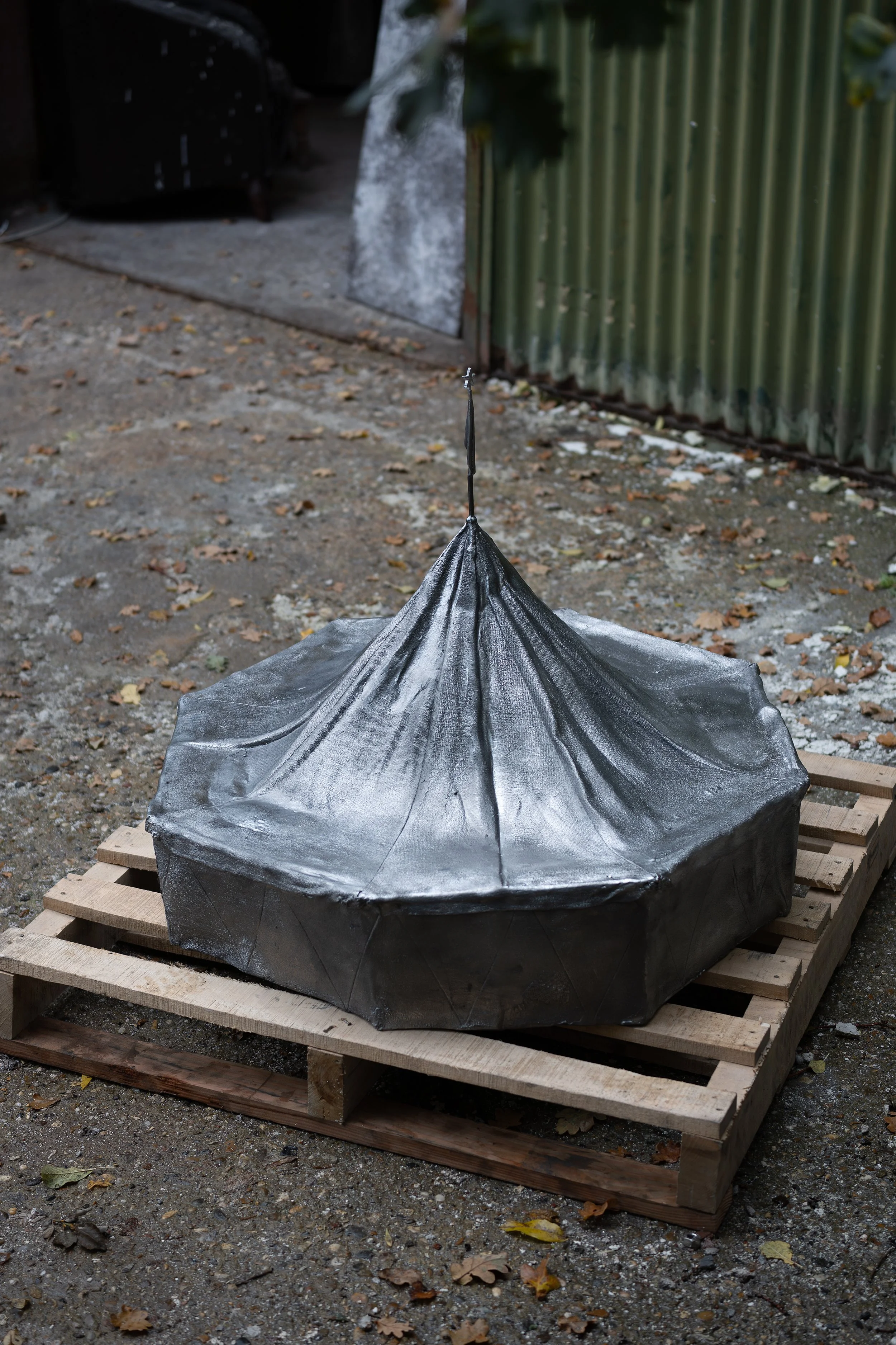 A large umbrella painted in metallic silver, resting on a wooden pallet outdoors on a dirt ground with fallen leaves.