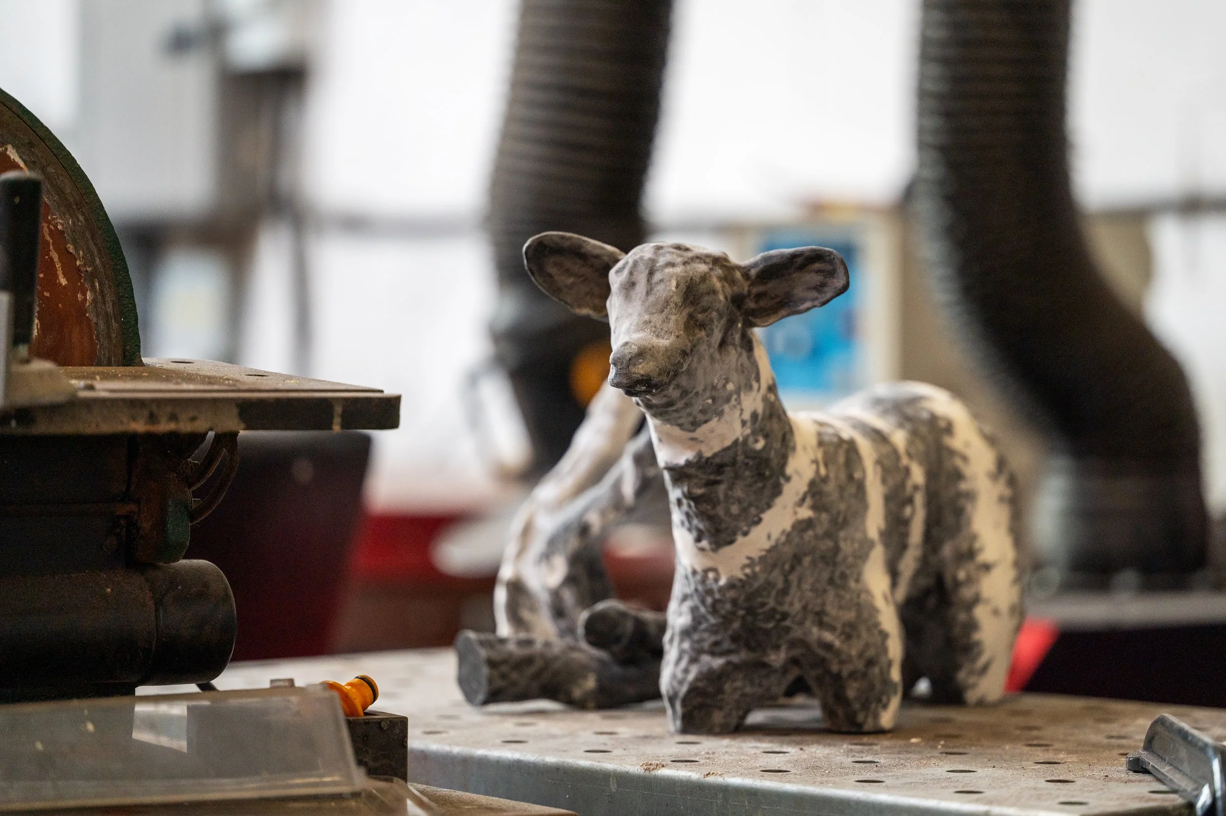 A close-up of a black and white speckled dog sculpture on a workbench in a workshop, with tools and equipment around.