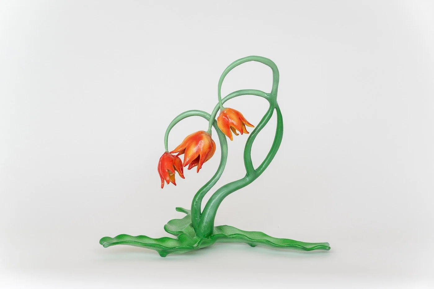 Glass sculpture of three orange and red flowers with green leaves and stems against a plain white background.