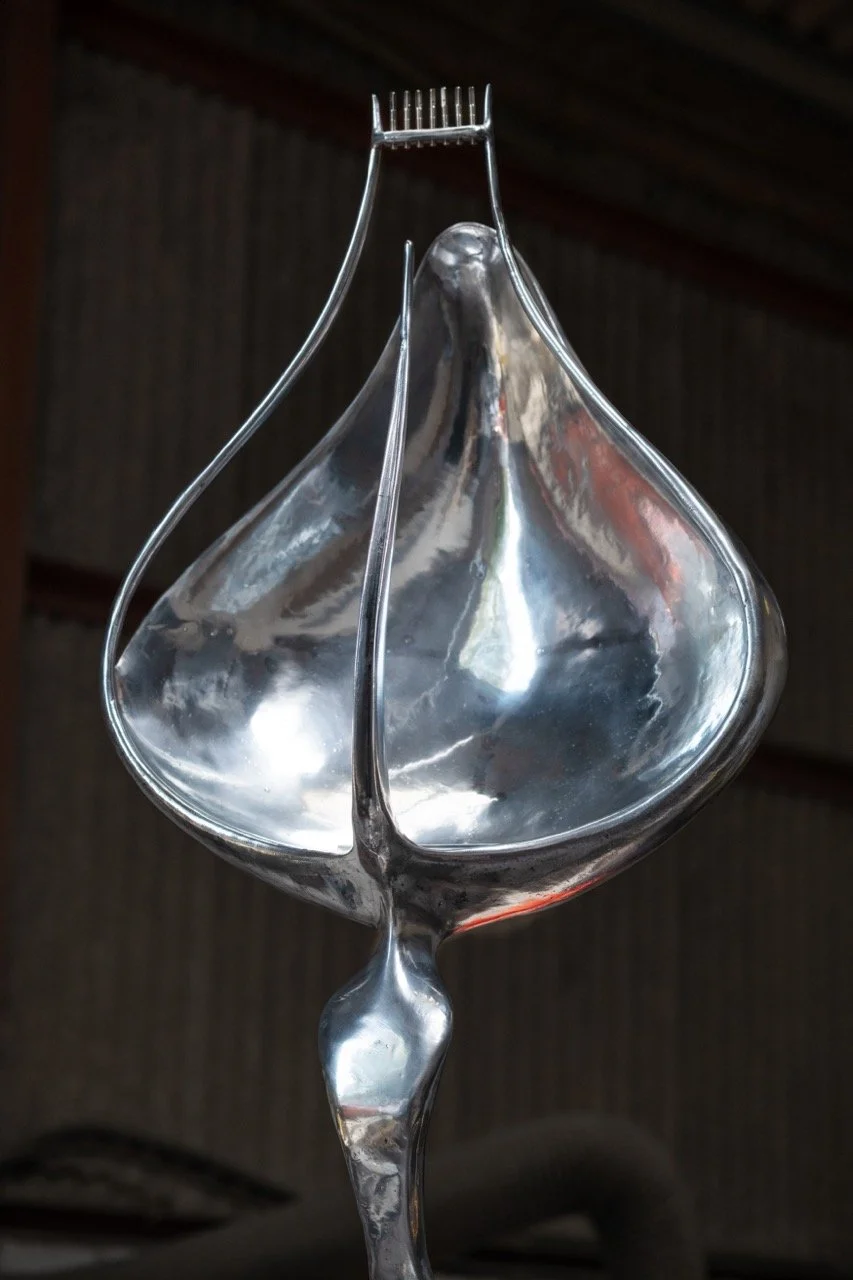 A silver decorative object resembling a stylized leaf or teardrop shape with a pointed top and a rounded bottom, mounted on a thin, curved silver rod.