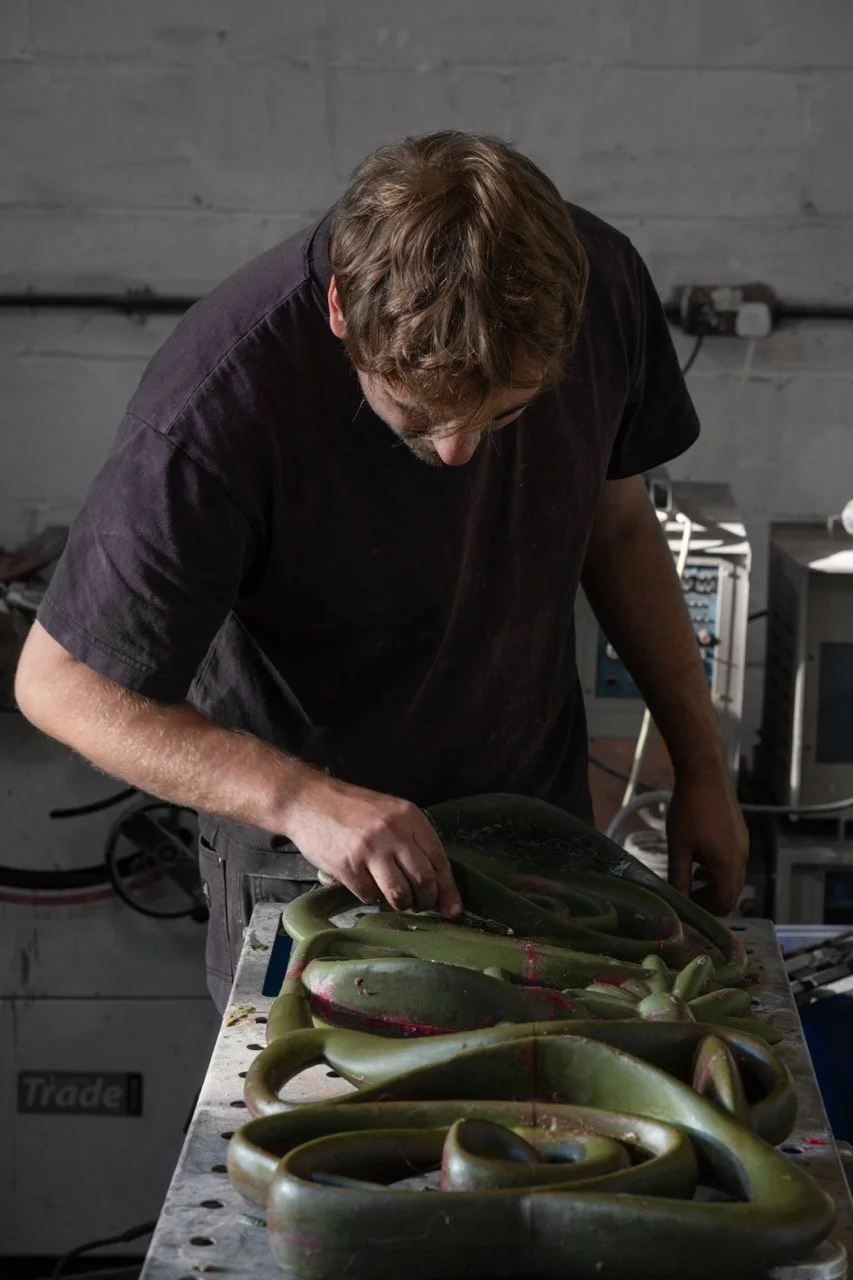 A man with brown hair and a black shirt working on a large, twisted green sculpture or artwork on a workbench.