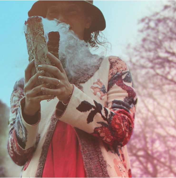 A person wearing a hat and floral-patterned sweater, holding an ice cream cone with smoke or vapor emanating from their face, in an outdoor setting with trees in the background.