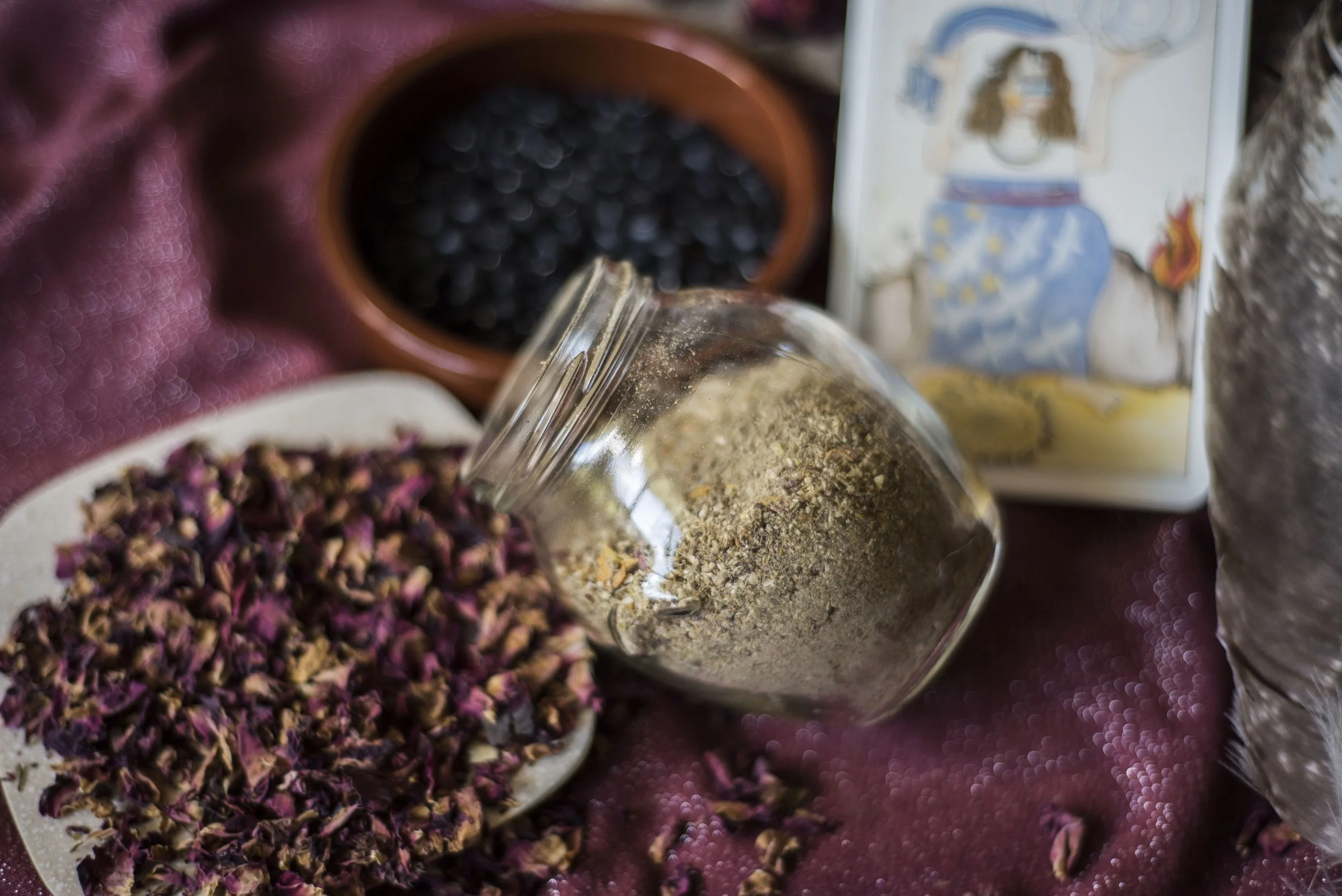 Spilled dried rosebuds, a glass jar of ground herbs, a bowl of dried blackberries, and a tarot card on a glittery purple surface.