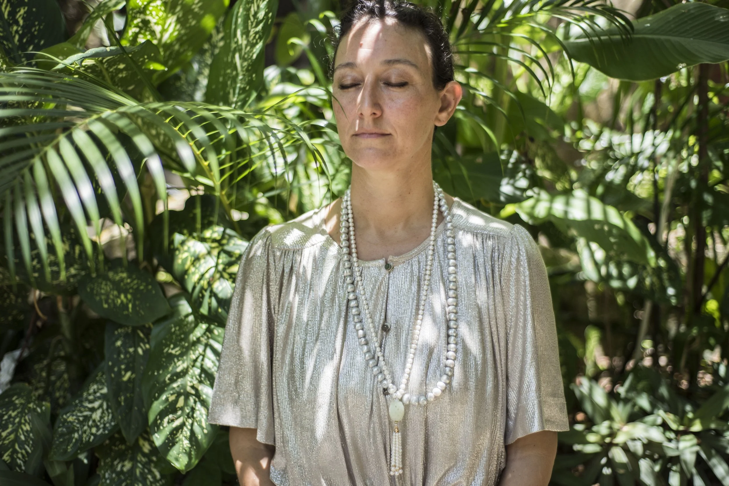 A woman with dark hair and closed eyes, wearing a shiny silver top and multiple pearl necklaces, stands among lush green tropical plants.