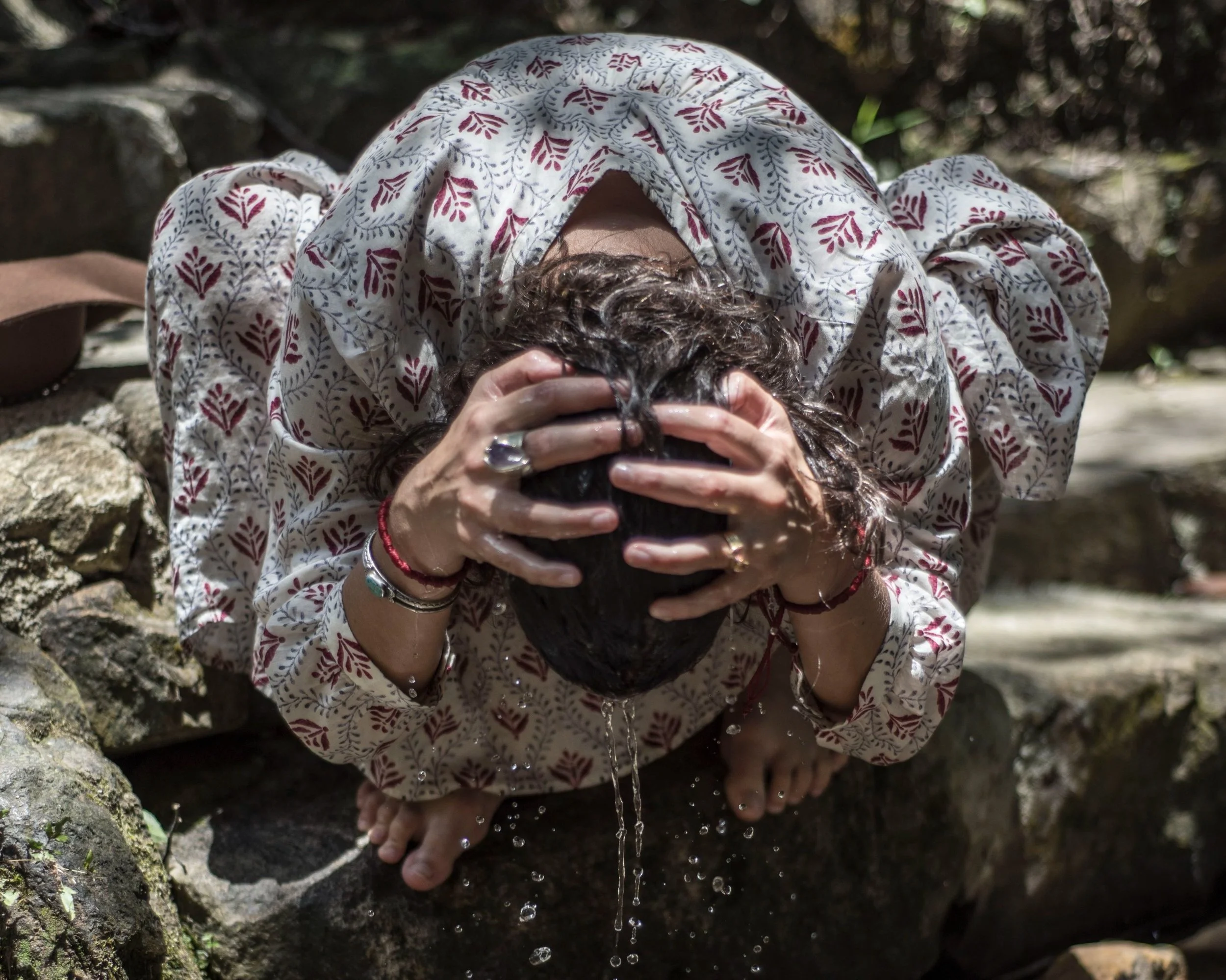 Woman crouching on a rock, washing her face with water, holding her head with both hands, wearing a patterned blouse, with water dripping from her face.