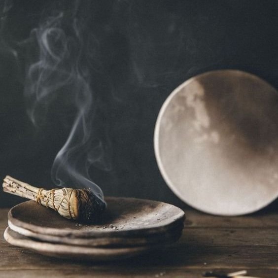 A lit incense cone on a stack of stones with smoke rising, and a round, reflective object in the background.