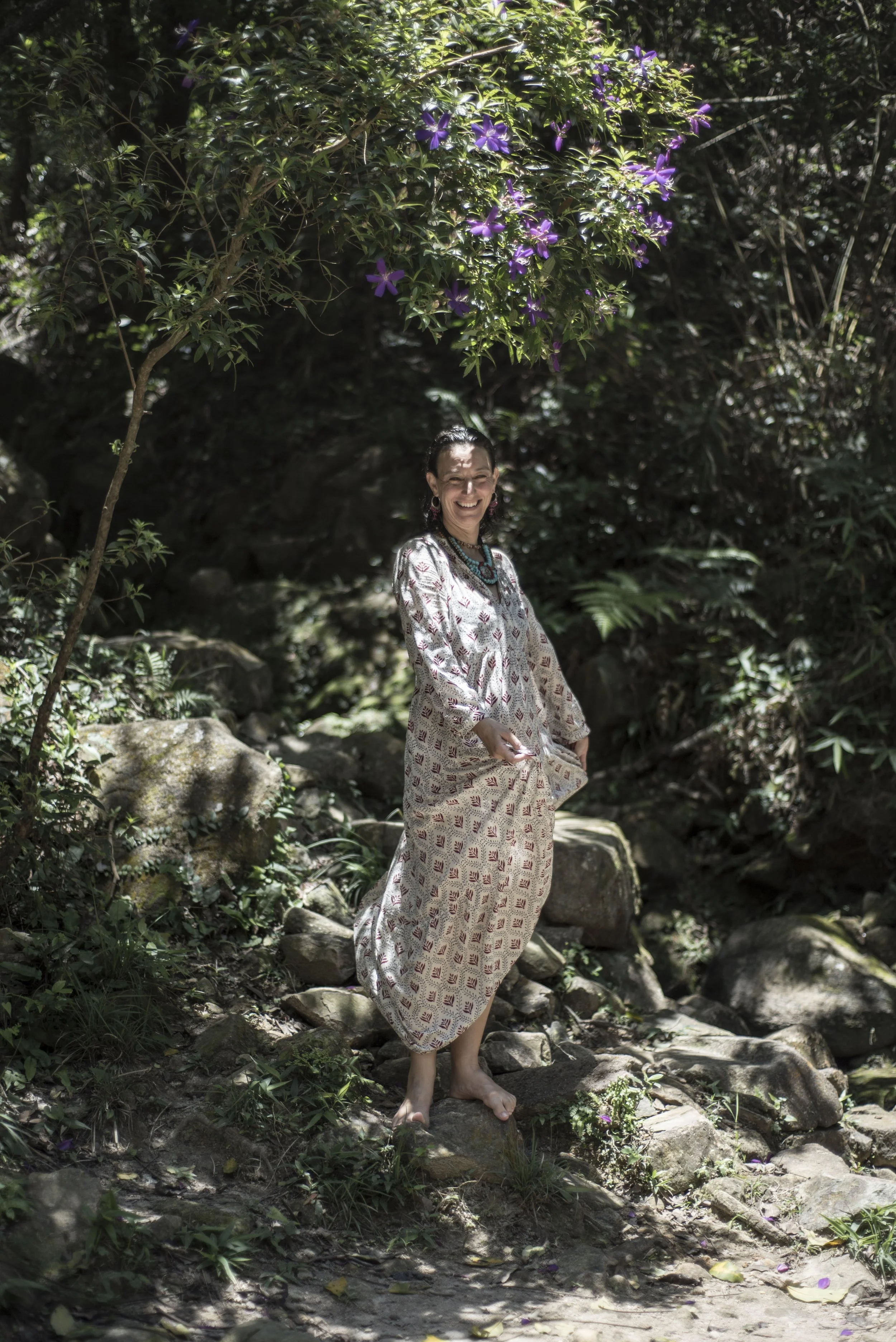 A woman smiling and holding a small object in a natural outdoor setting with rocks, greenery, and purple flowers overhead.