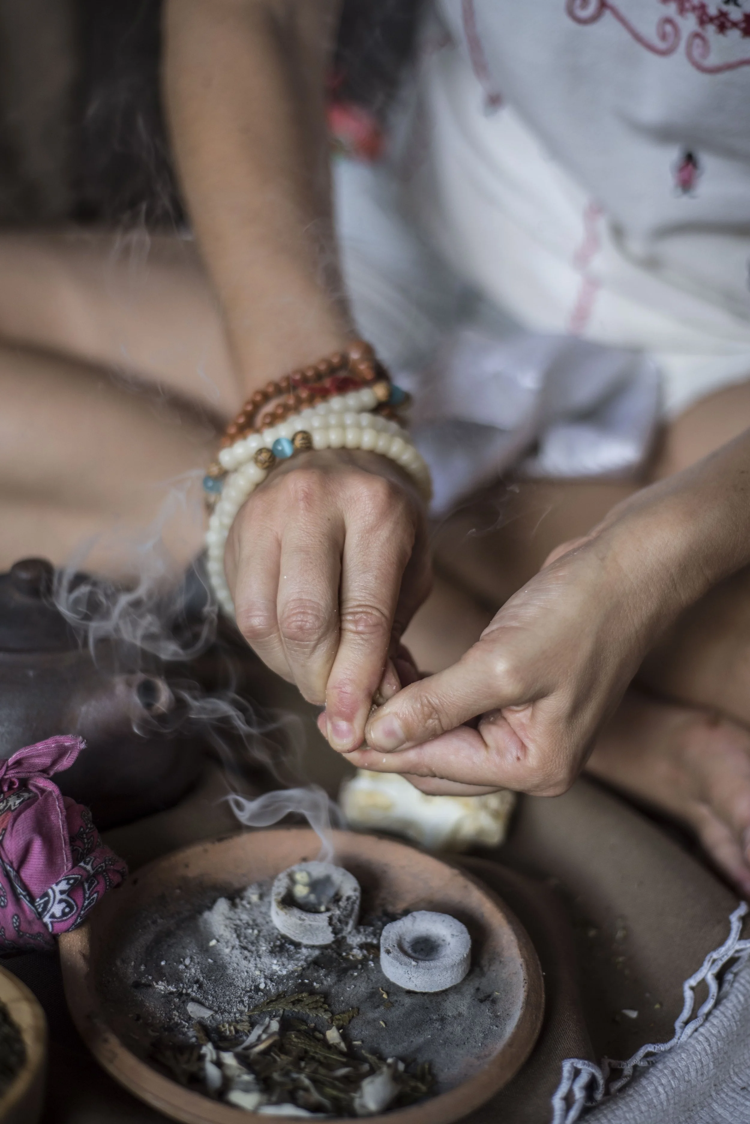 Person wearing multiple beaded bracelets, sitting cross-legged, lighting incense sticks over an incense holder with ash and burned incense remains.