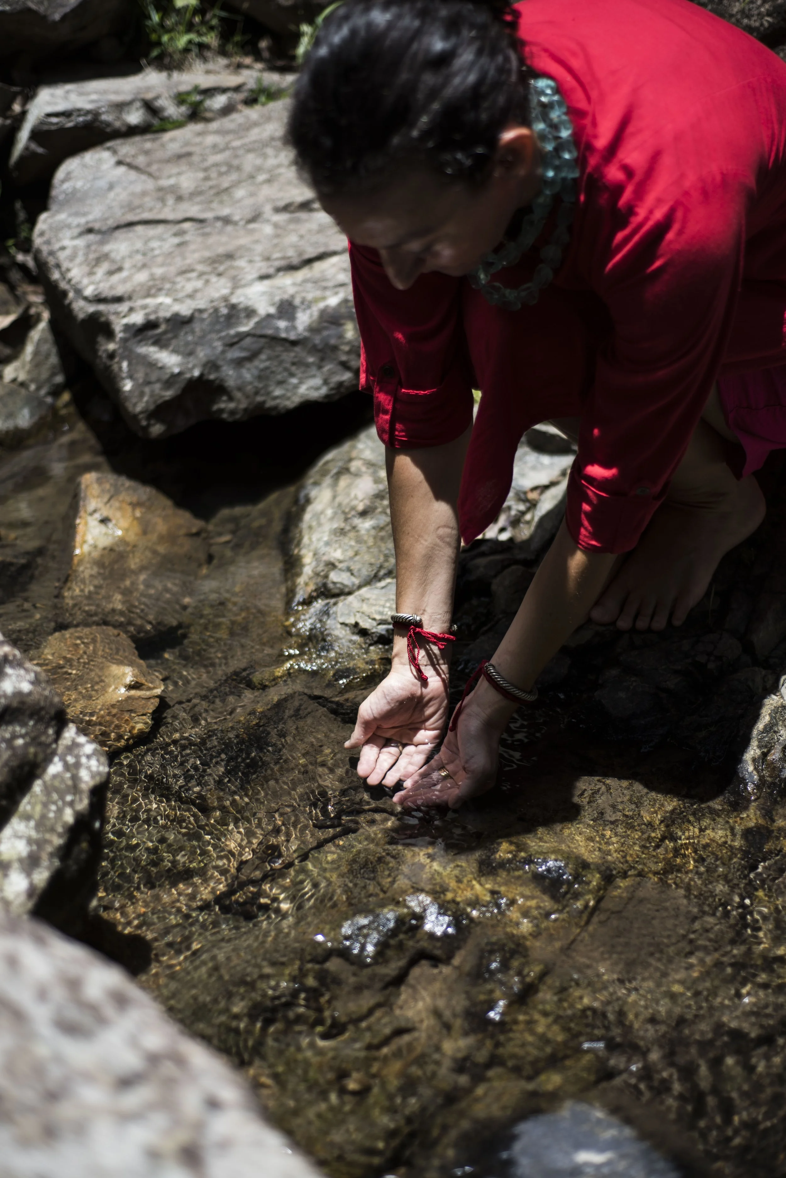 Person with short dark hair wearing a red shirt, turquoise necklace, and bracelets, crouching by a small stream with rocks, touching the water with both hands.