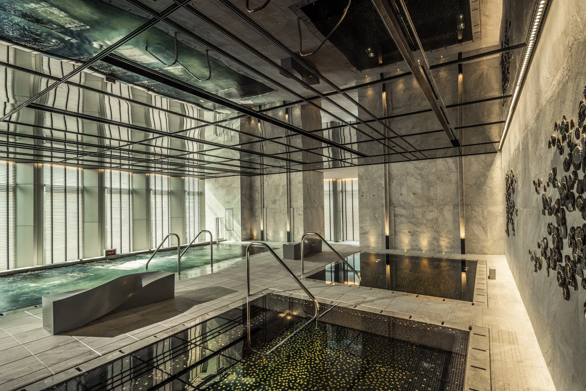 Modern indoor spa with two hot tubs, one with black mosaic tiles and the other with clear water, stainless steel handrails, stone flooring, marble walls, large windows with vertical blinds, and artistic wall decor.