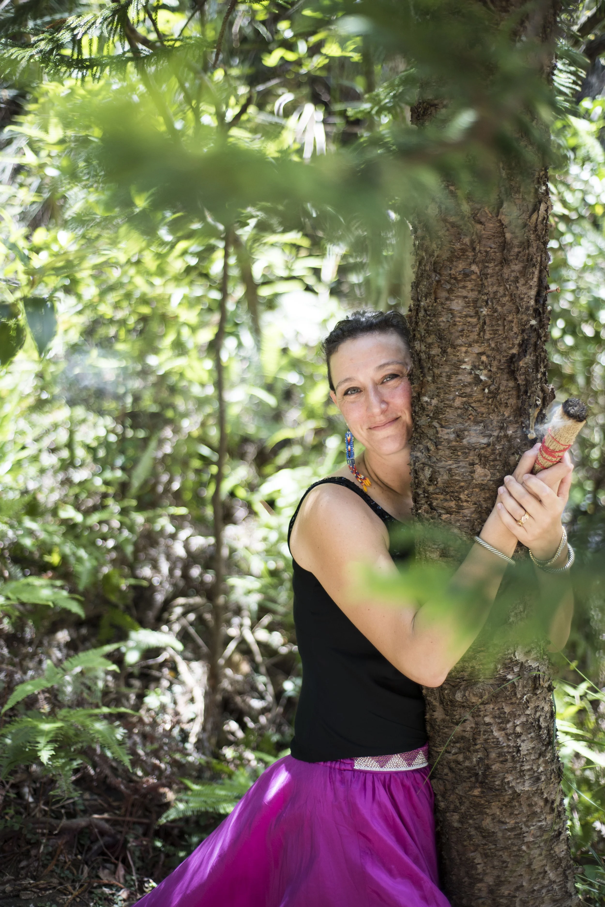 A woman with dark hair, wearing a black top, a purple skirt, colorful jewelry, and makeup, is hugging a tree in a lush green forest, smiling at the camera.