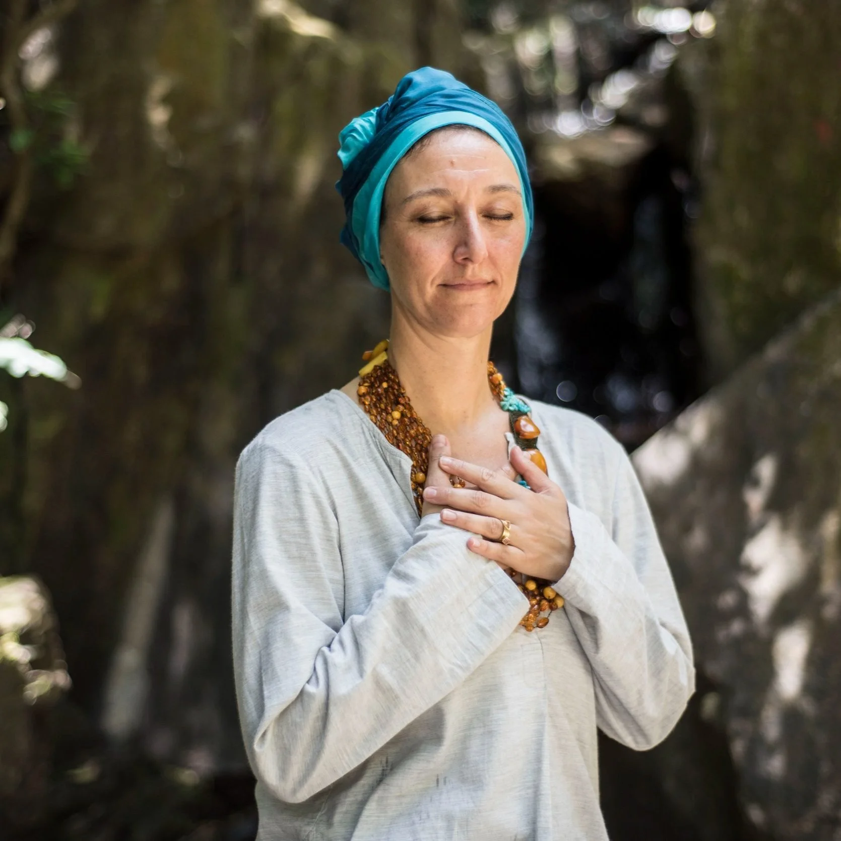 A woman with a blue headscarf and light gray shirt, her eyes closed, touching her chest with her right hand, standing in front of a waterfall in a forest.