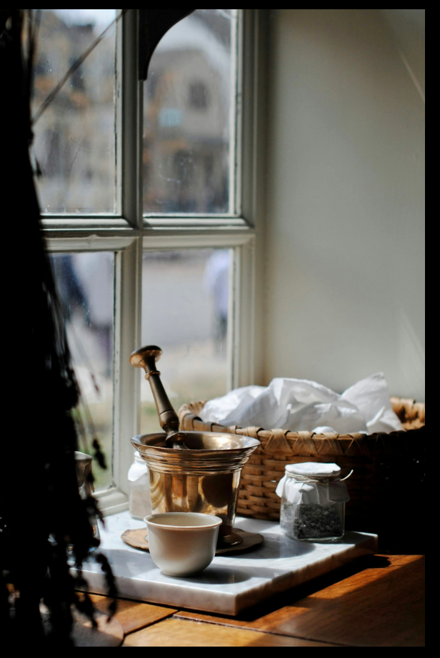 Kitchen windowsill with a white bowl, a mortar and pestle, a jar of herbs, and a wicker basket filled with white cloth, illuminated by natural light.