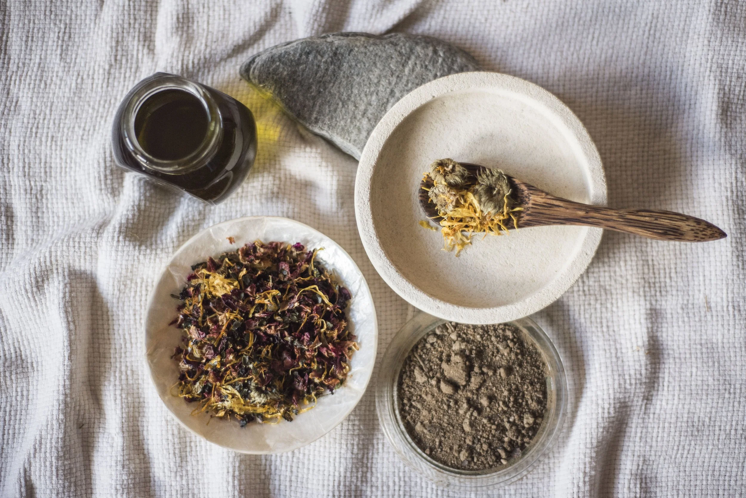 Various herbal powders, dried herbs, and tea ingredients arranged on plates and bowls on a textured beige cloth.