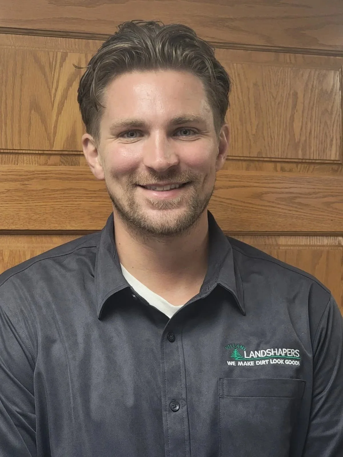A smiling man wearing a dark gray work shirt with the logo of Villani Landshapers and the slogan "We Make Dirt Look Good!" is standing in front of a wooden wall.
