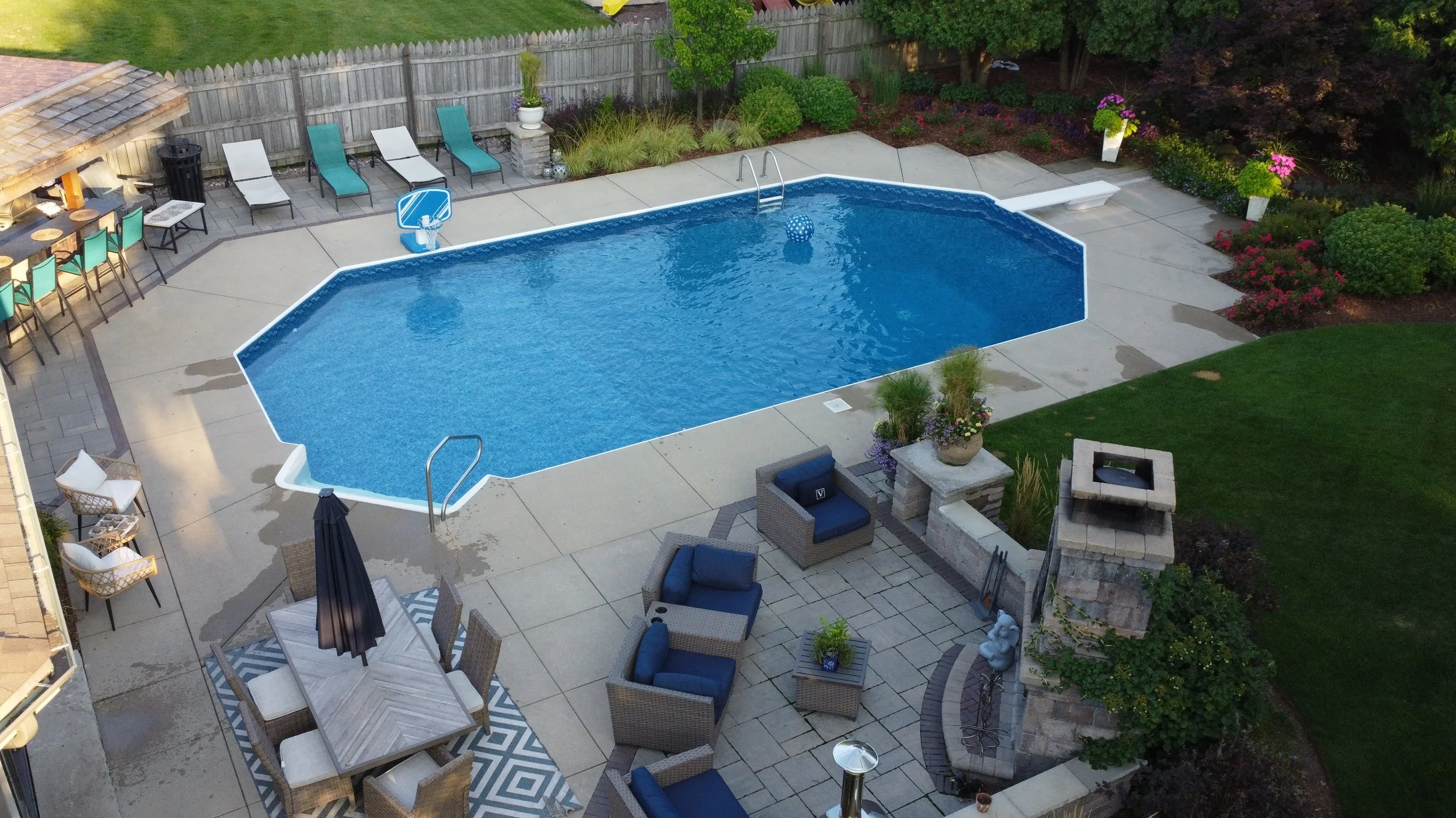A backyard with a swimming pool, poolside chairs, outdoor dining table with chairs, and a fire pit area surrounded by lush greenery and colorful flowers.