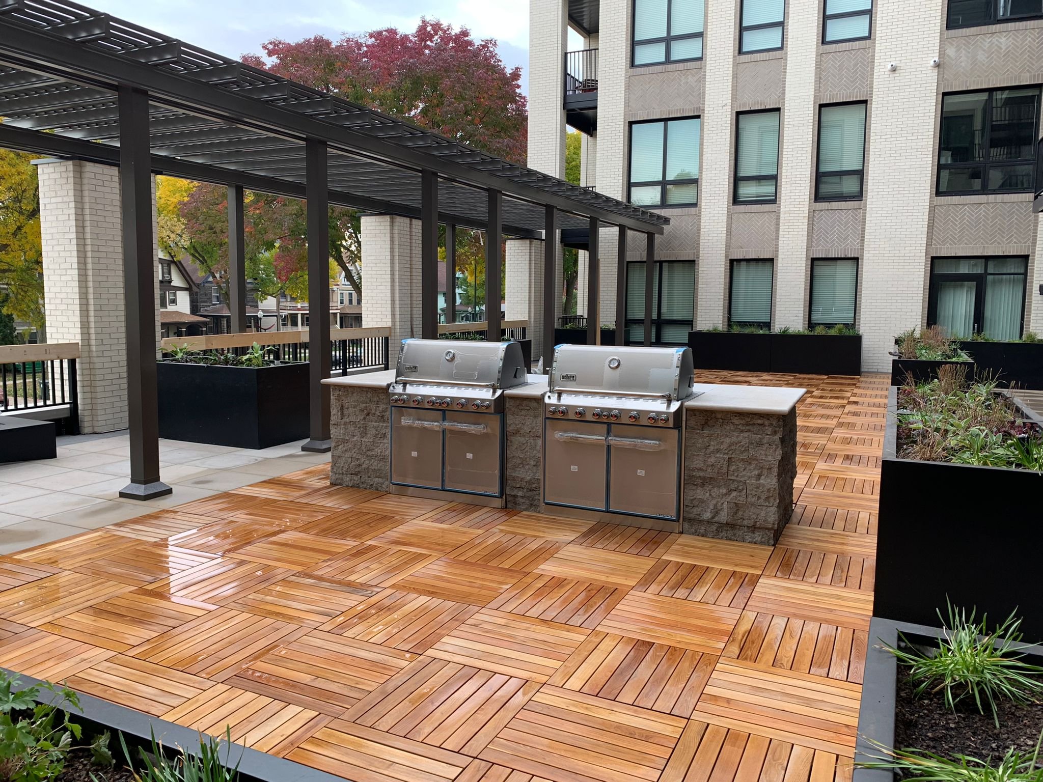 Outdoor balcony with two stainless steel gas grills, wooden tile flooring, black planters with plants, and a building with multiple windows in the background.