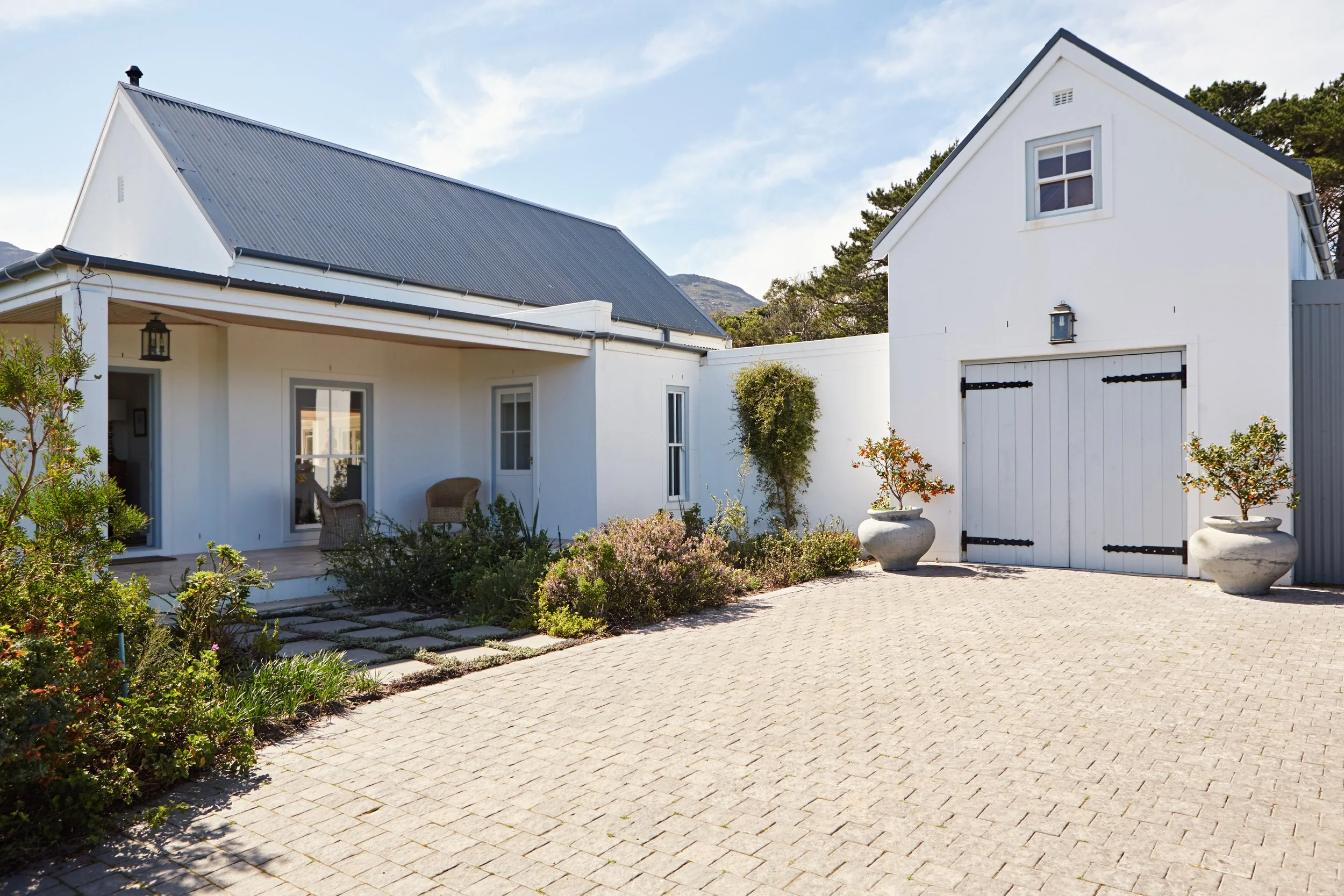 White house with a pitched gray roof, attached to a garage with a similar roof, surrounded by a stone-paved driveway and landscaped plants in large pots and bushes, under a partly cloudy sky.