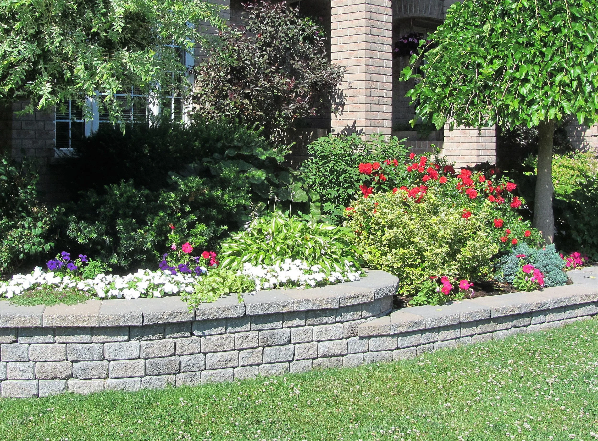 A well-maintained front yard with colorful flowers, green bushes, and trees in front of a brick house.