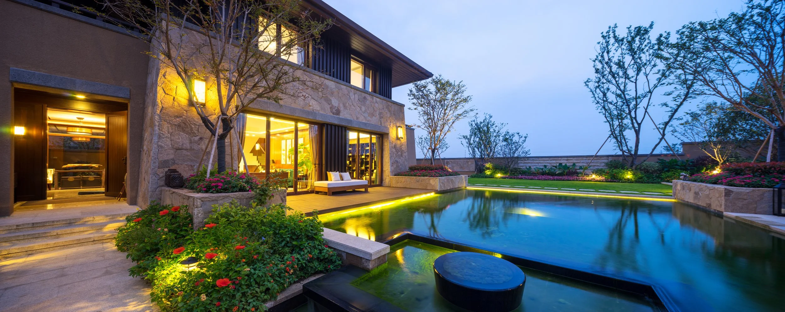 Luxury modern house with a swimming pool, outdoor seating, landscaped garden with flowers and trees at dusk