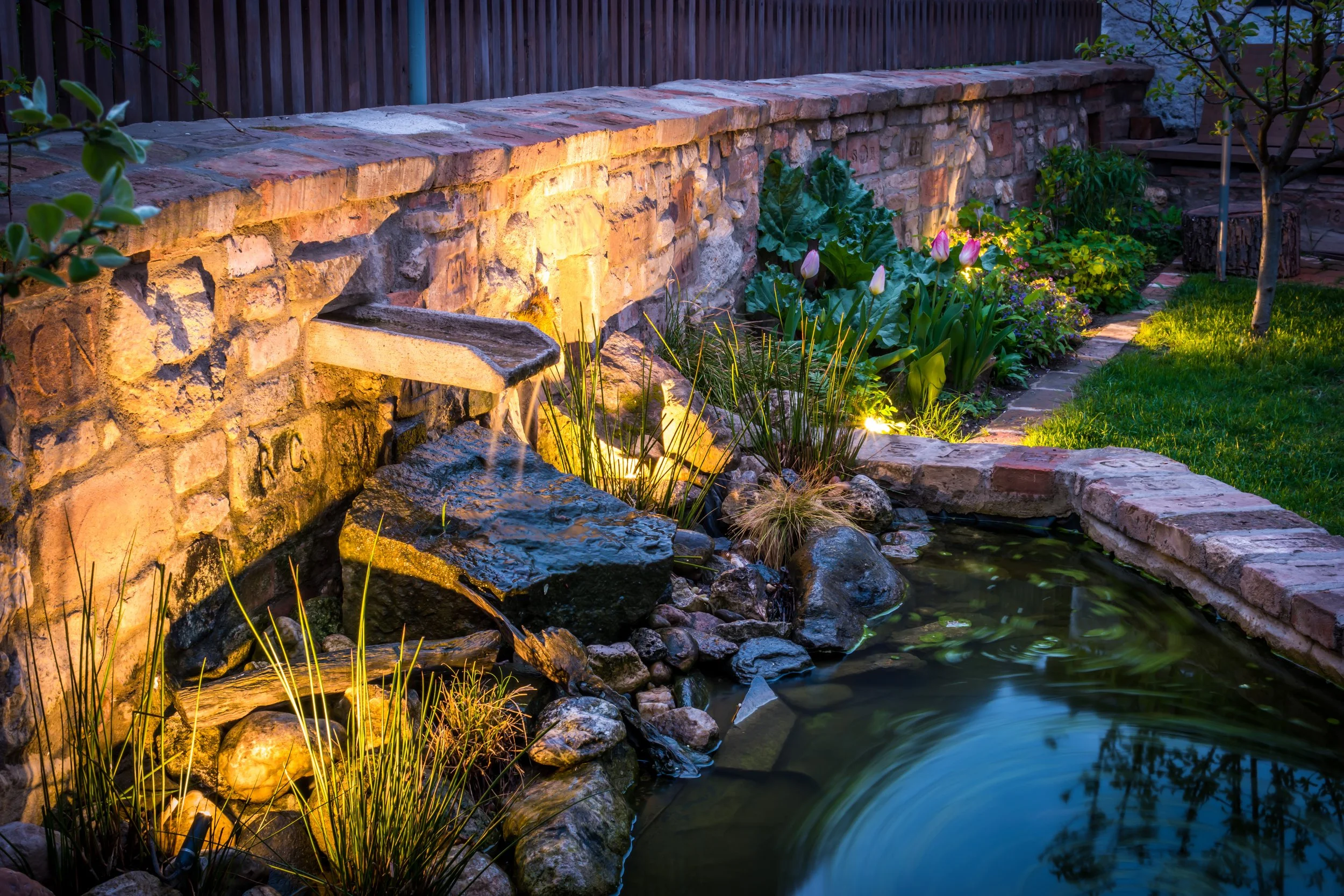 Decorative backyard pond with waterfall, illuminated by outdoor lighting, bordered by rocks, plants, tulips, and a brick and stone retaining wall at dusk.