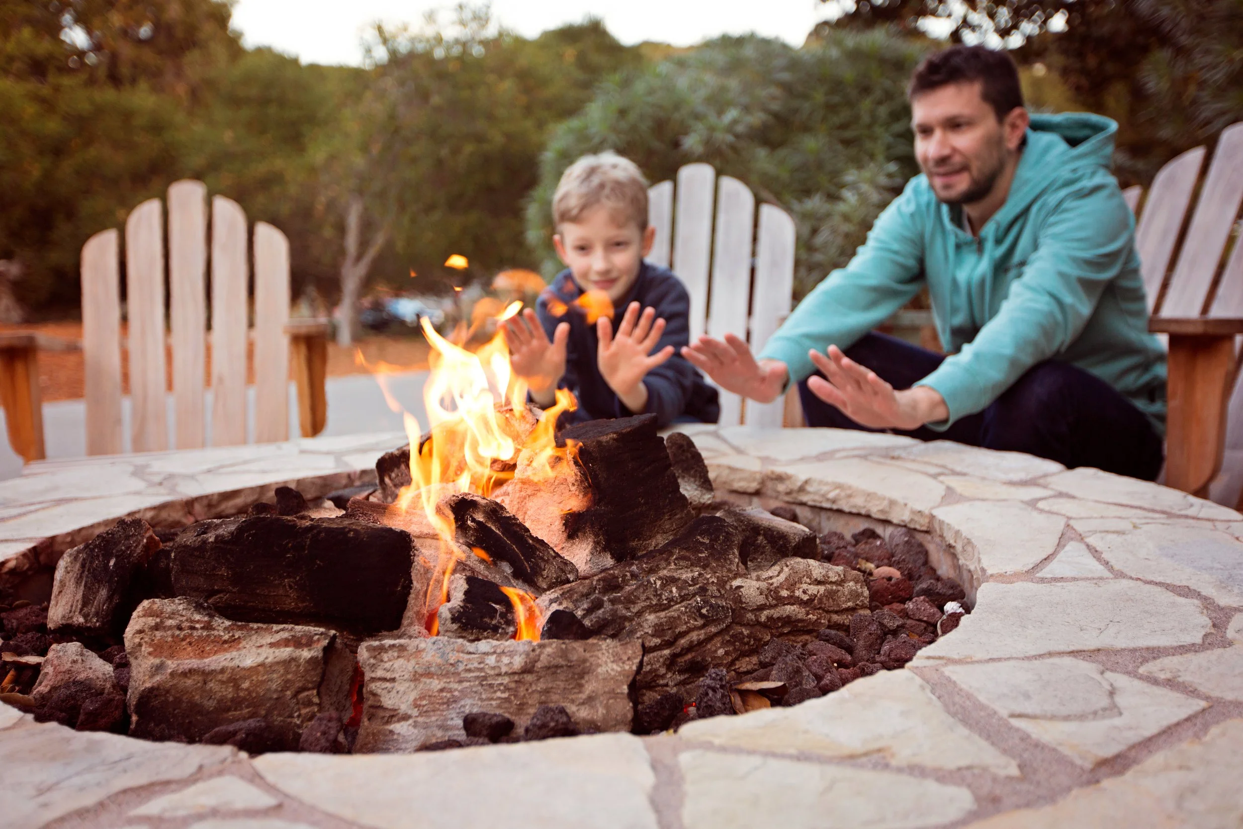 A man and boy sitting at a stone fire pit, warming their hands over a campfire outdoors, with trees and a fence in the background.