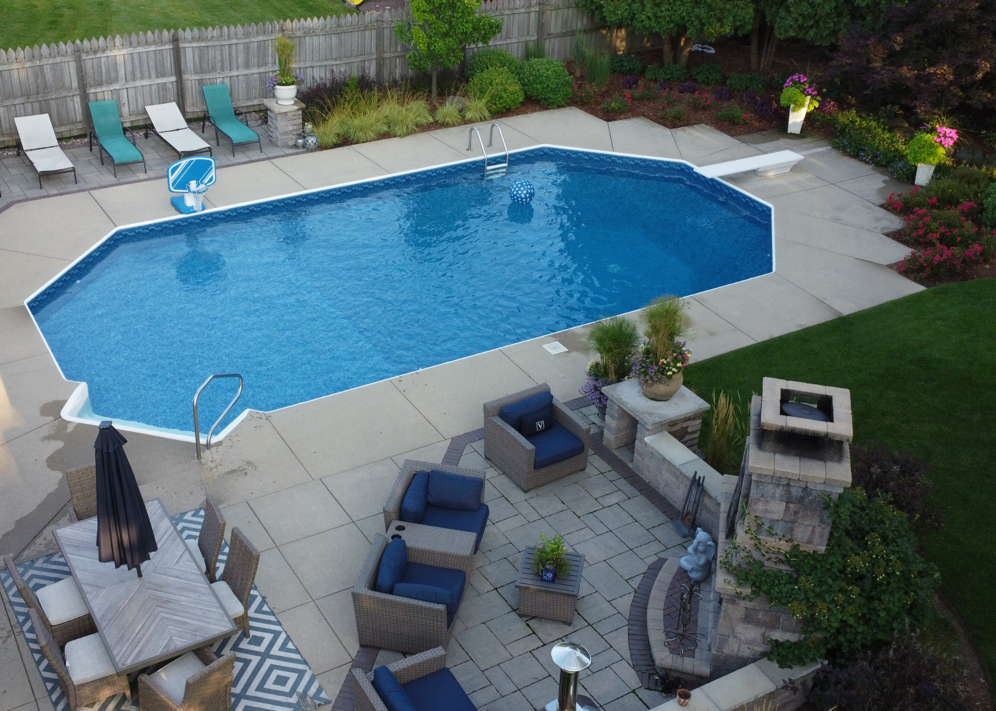 A backyard swimming pool surrounded by patio furniture, lounge chairs, and landscaped garden with colorful flowers and greenery.