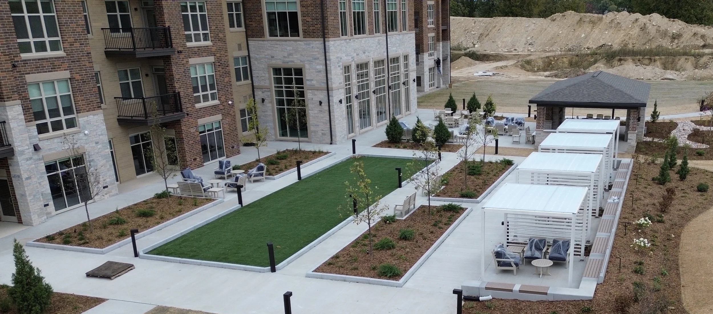 A modern apartment complex outdoor courtyard with trees, seating areas, and a grassy area, adjacent to a multi-story building, with a covered pavilion and landscaped pathways.