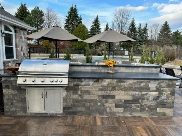 Outdoor grill island with two umbrellas, decorative flowers, and a backyard view of trees and a tennis court.