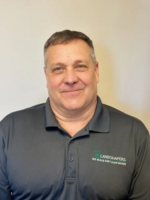 A smiling man in a dark gray polo shirt with logo and text, standing against an off-white wall.