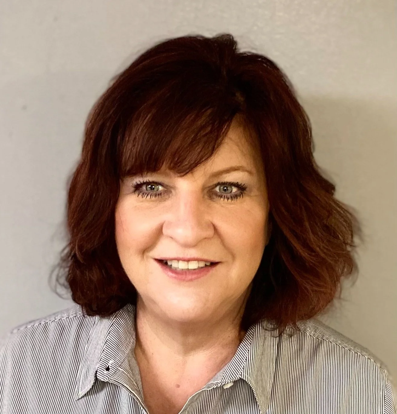 A woman with shoulder-length red hair and blue eyes smiling at the camera, wearing a light-colored striped shirt, standing against a plain light gray wall.