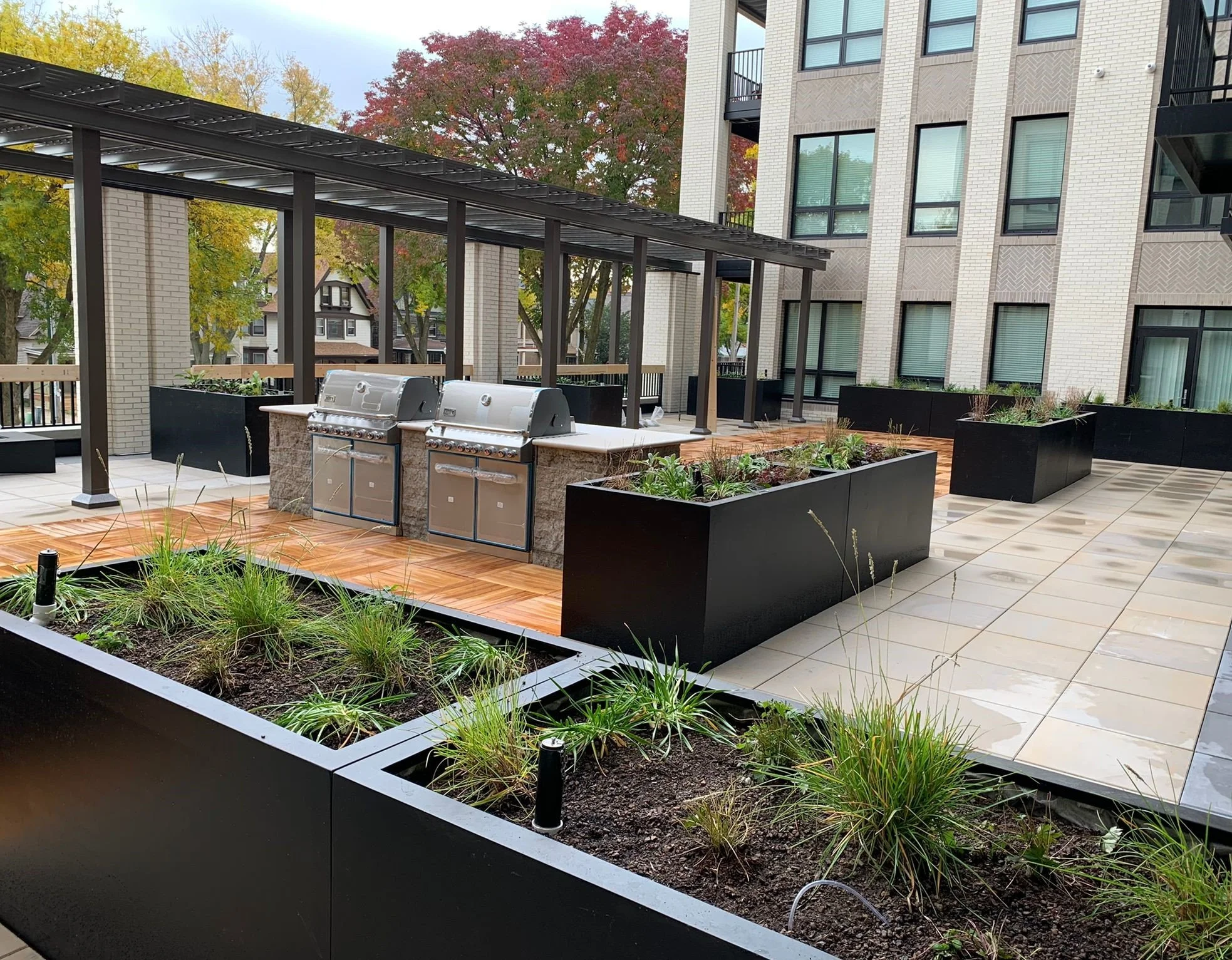 Modern outdoor balcony with built-in grills, black rectangular planters with greenery, wooden and tiled flooring, and a metal pergola with glass roof panels at an apartment complex during autumn.