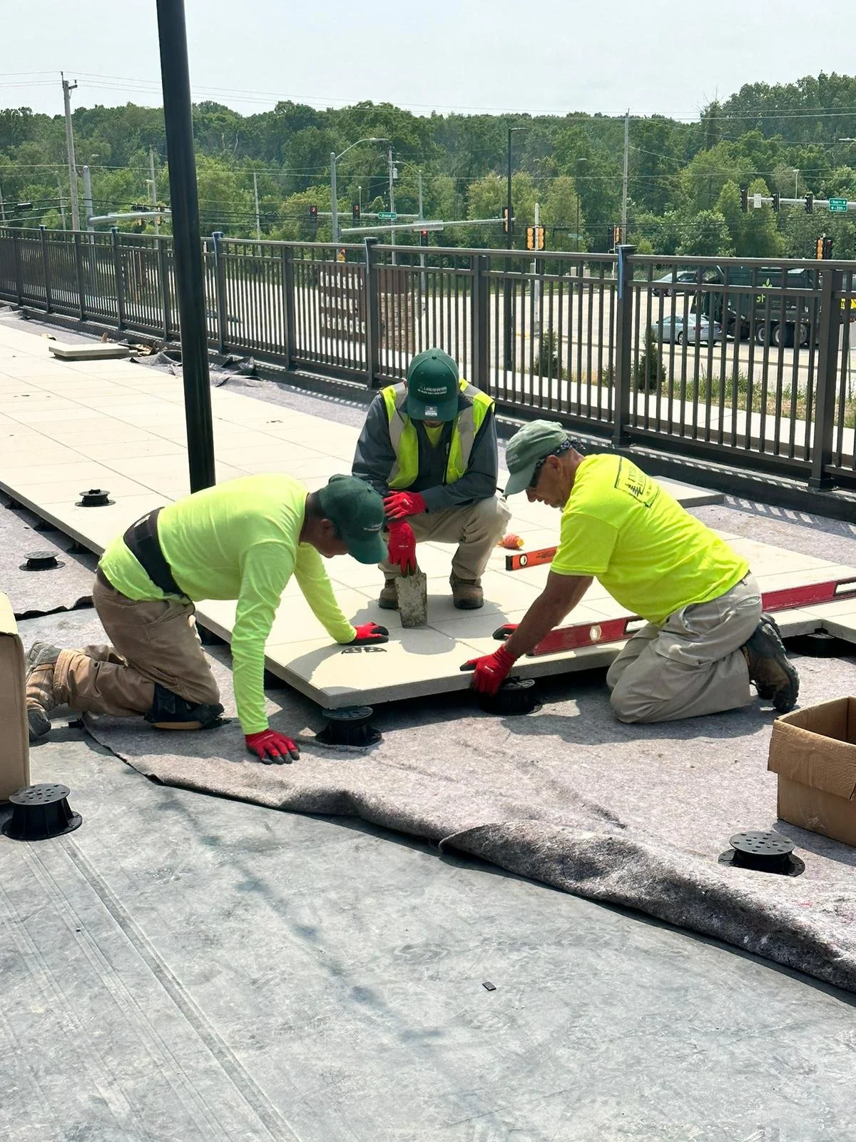 Three construction workers installing large tiles on a rooftop, wearing safety gear and working with tools.
