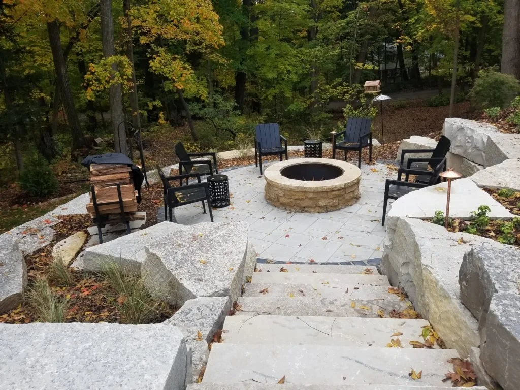 Outdoor fire pit patio surrounded by black chairs, stone steps, and large rocks, with a wooded background and fall leaves.