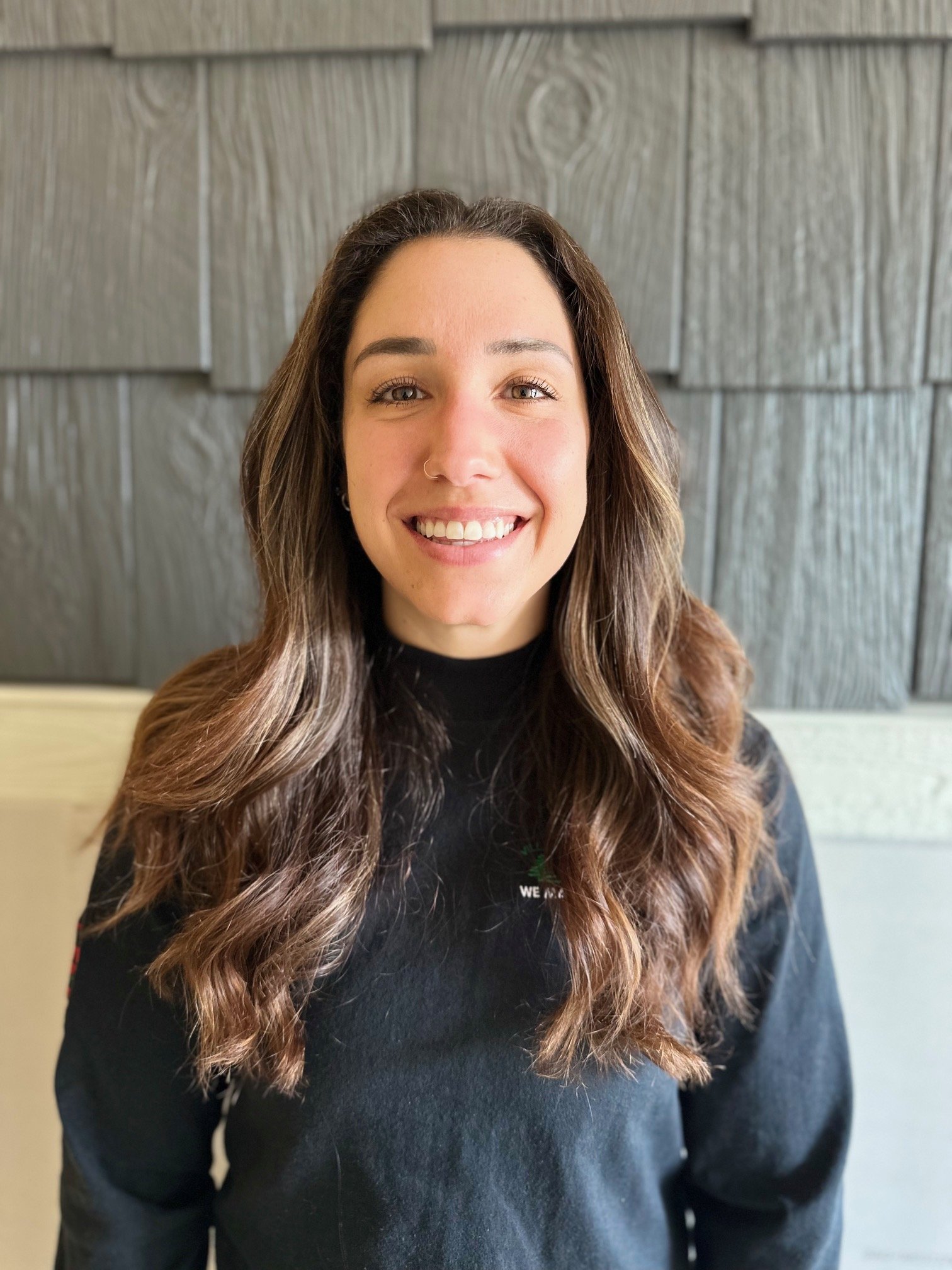 A young woman with long, wavy brown hair, smiling, wearing a black shirt, standing in front of a wall with gray wooden panels.