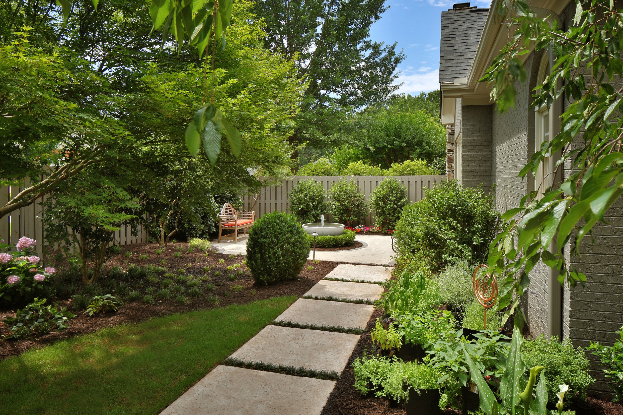 Residential backyard garden with a stone pathway, lush green trees, shrubs, flowering plants, a decorative water fountain, and a wooden bench on a concrete slab.