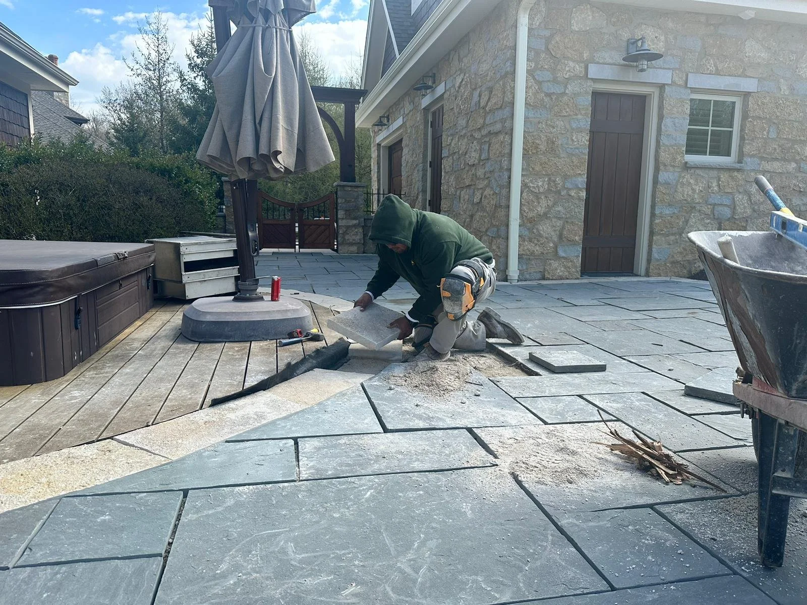 Person installing or repairing stone tiles on a patio, kneeling down with tools nearby, with a house and outdoor furniture in the background.